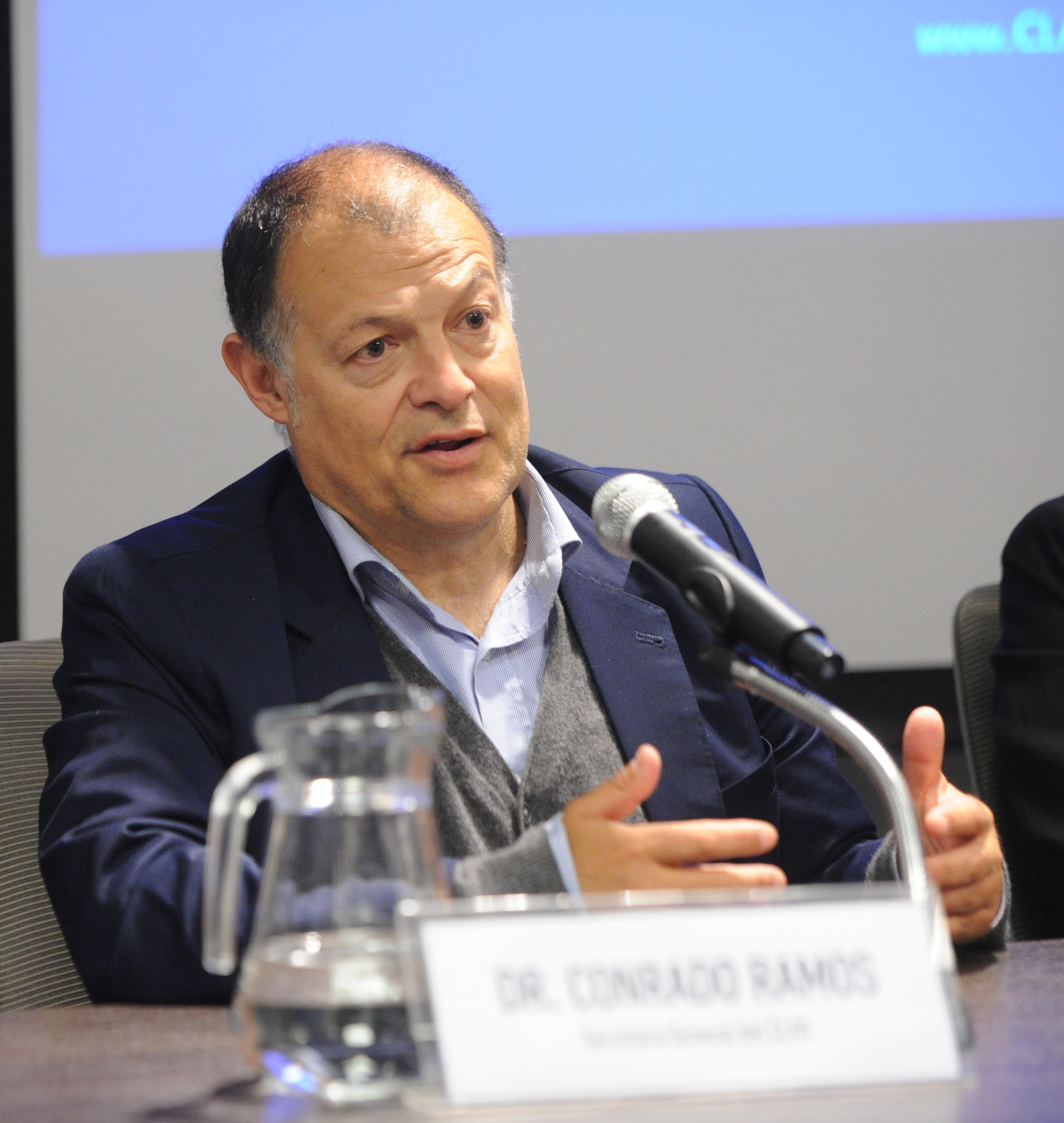 Secretario general del Centro Latinoamericano de Administración para el Desarrollo diserta sentado en mesa con micrófono.