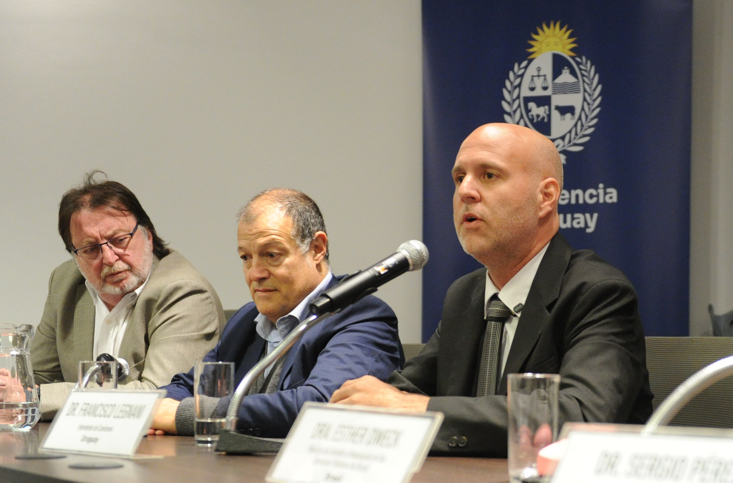 Intendente de Canelones diserta sentado en mesa con micrófono, mientras dos oradores lo observan a su lado.