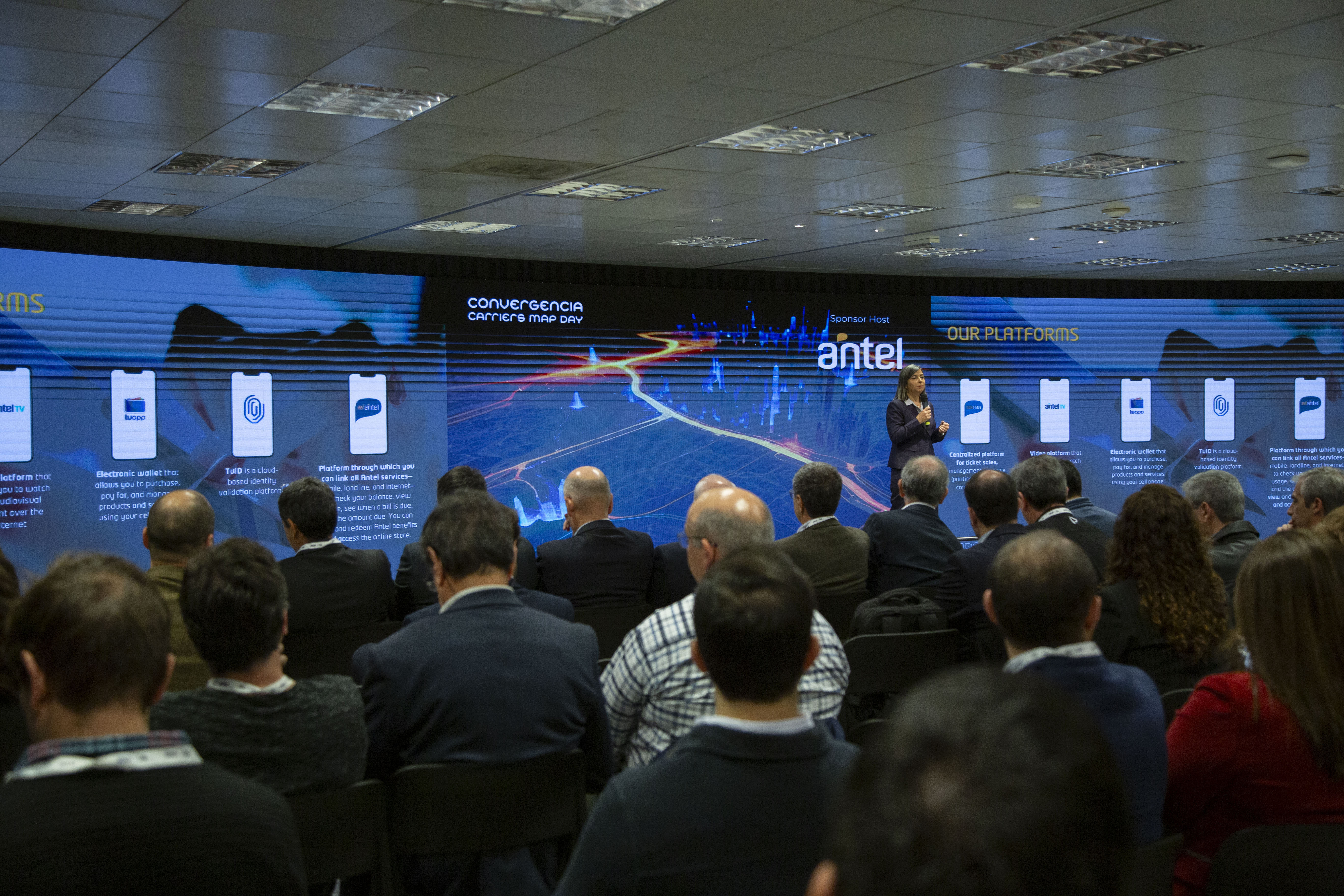 Plano general de los asistentes de espaldas escuchando la disertación de la gerenta general de Antel. Atrás se observa una pantalla con gráficos.