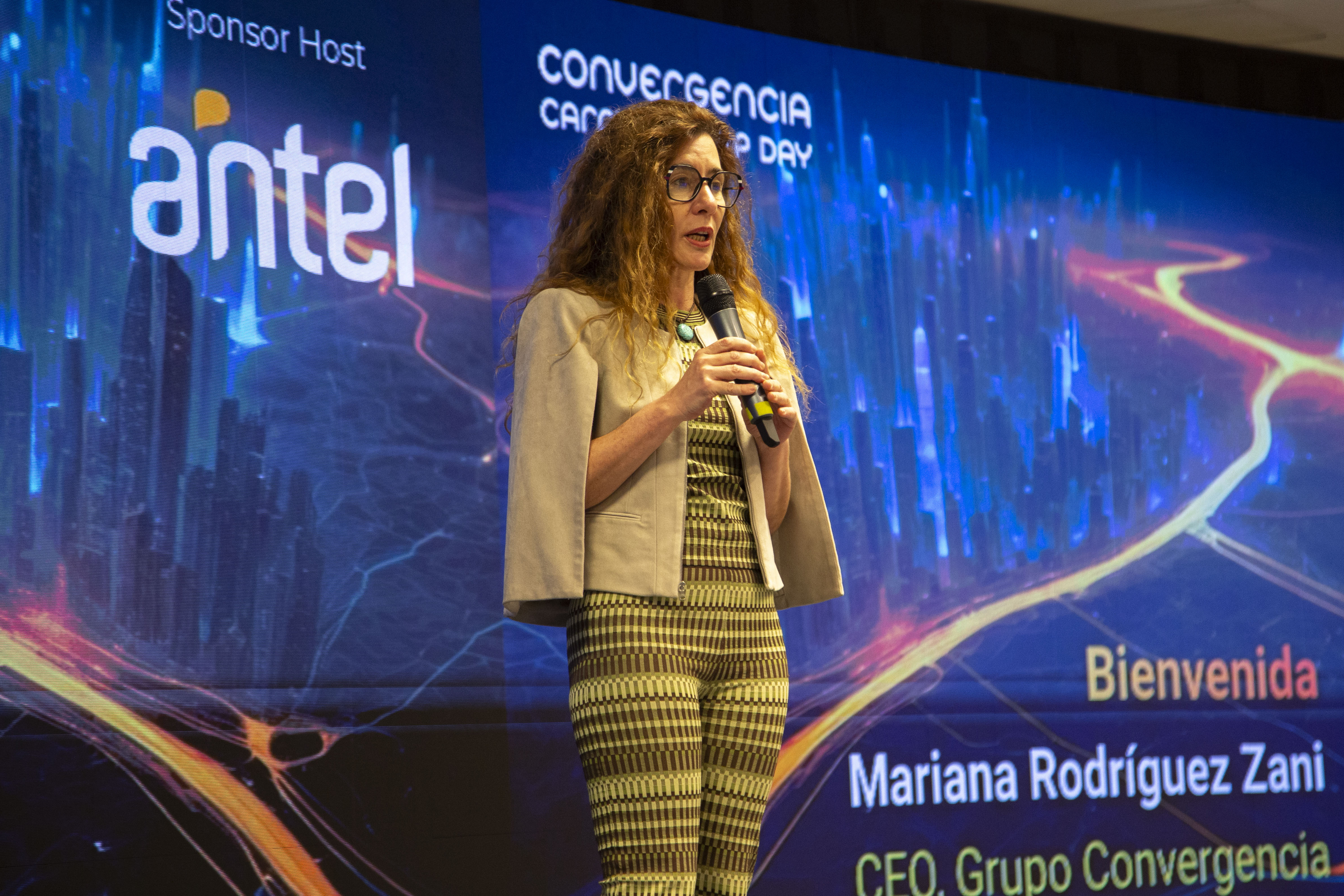 Primer plano de la CEO del Grupo Convergencia disertando de pie con micrófono en mano en el escenario. Atrás se observa una pantalla con gráficos.