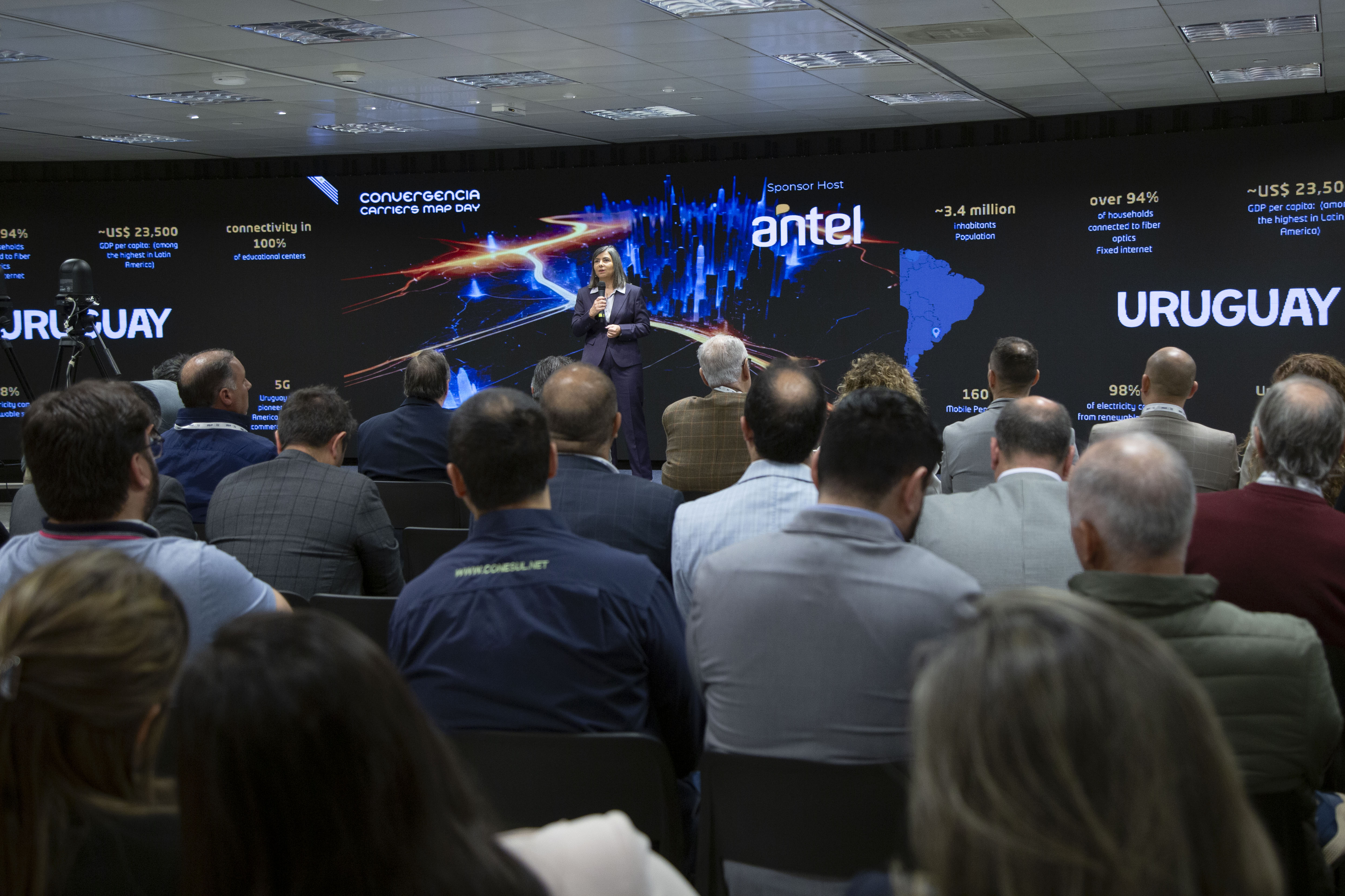 Plano general de los asistentes de espaldas escuchando la disertación de la gerenta general de Antel. Atrás se observa una pantalla con gráficos.