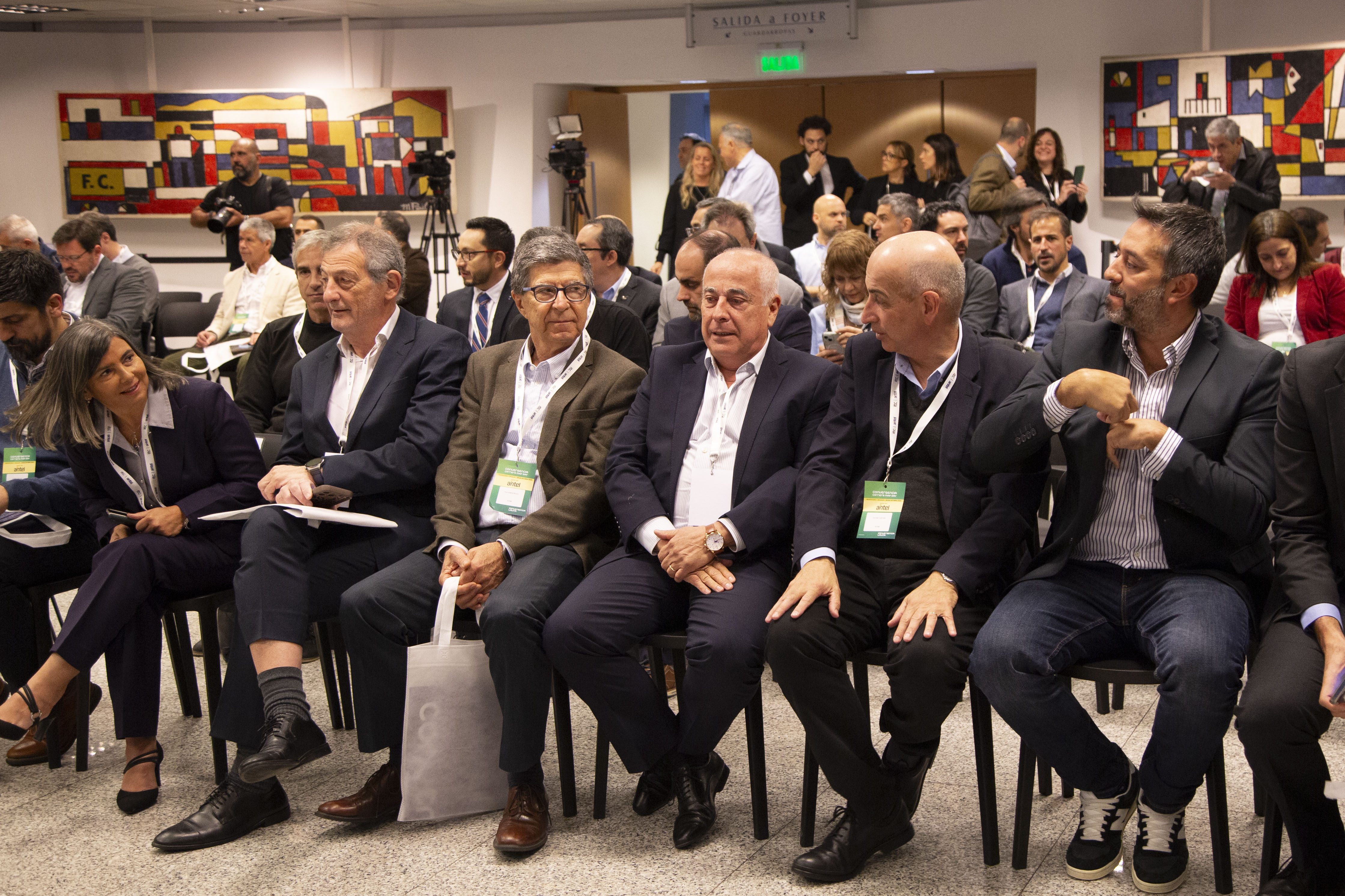 Plano general de un grupo de autoridades y participantes del evento dialogando sentados en primera fila del salón de actos.