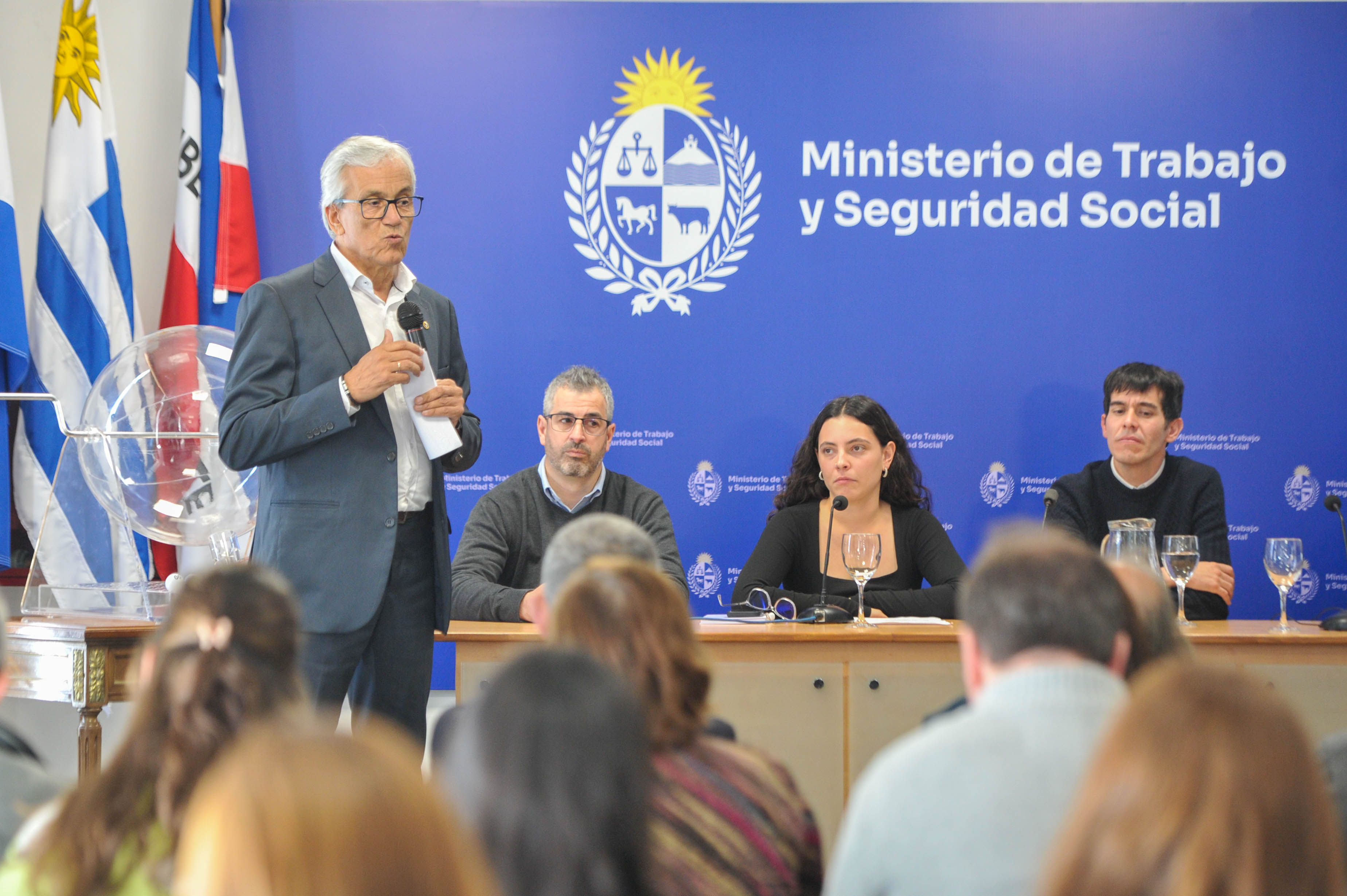 Ministro de Trabajo diserte de pie con micrófono en mano. Atrás están las autoridades sentadas en mesa principal observándolo.