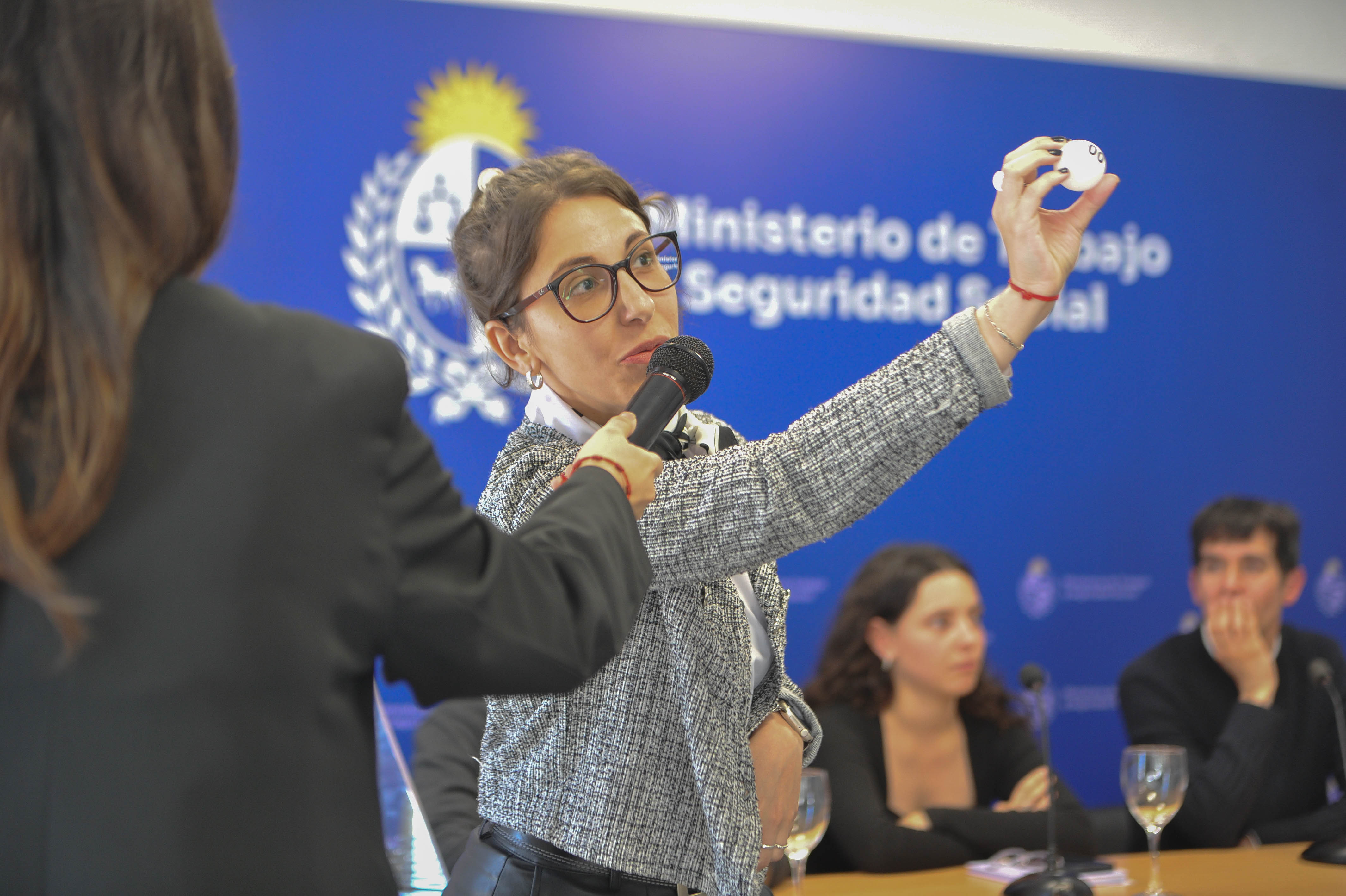 Una mujer con micrófono muestra una bola de sorteo blanca. Atrás se observa la mesa con las autoridades y cartel azul con logo del ministerio.