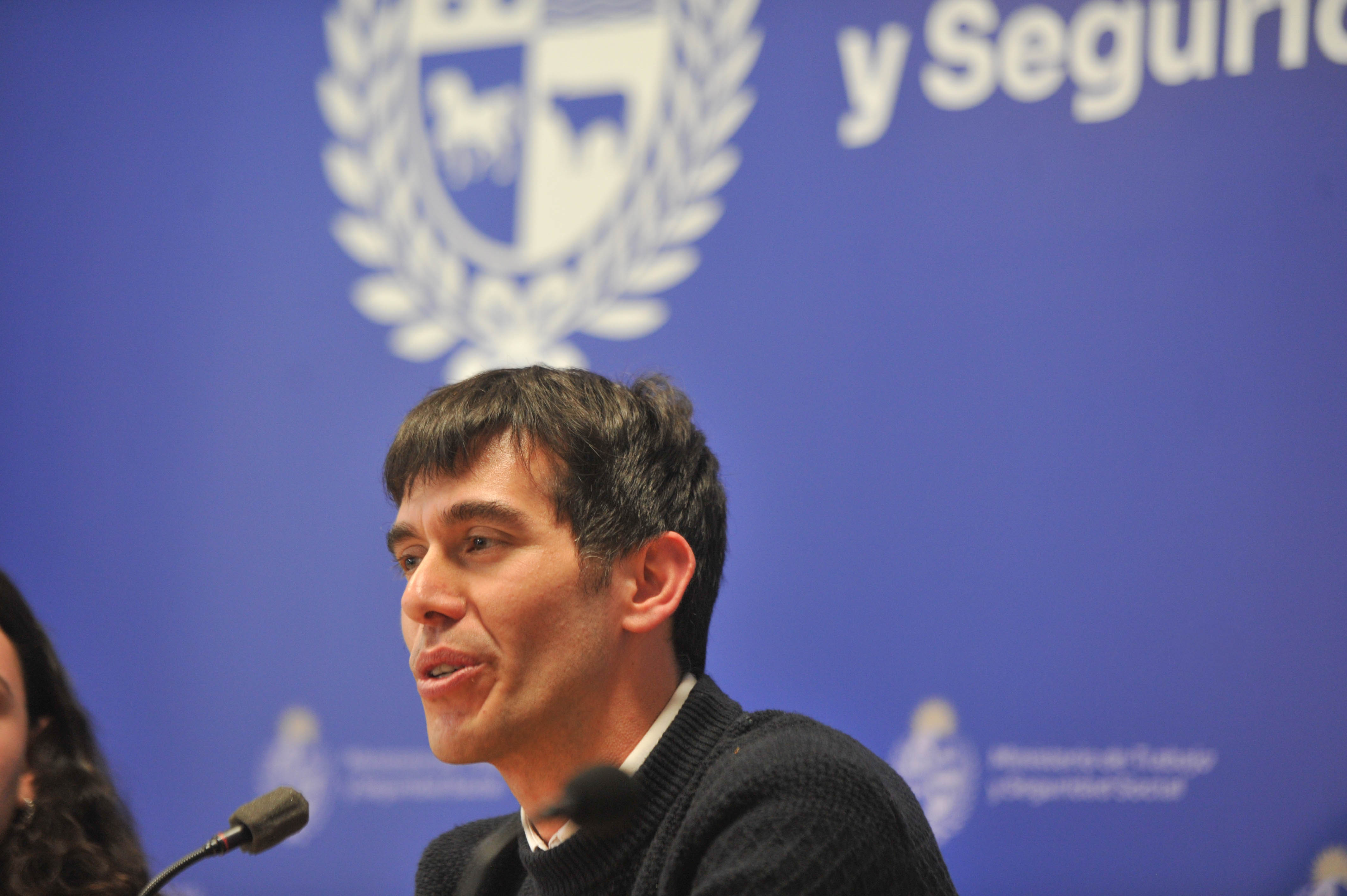 Director nacional de Trabajo diserta sentado en mesa con micrófono. Atrás se observa cartel azul con logo del ministerio.