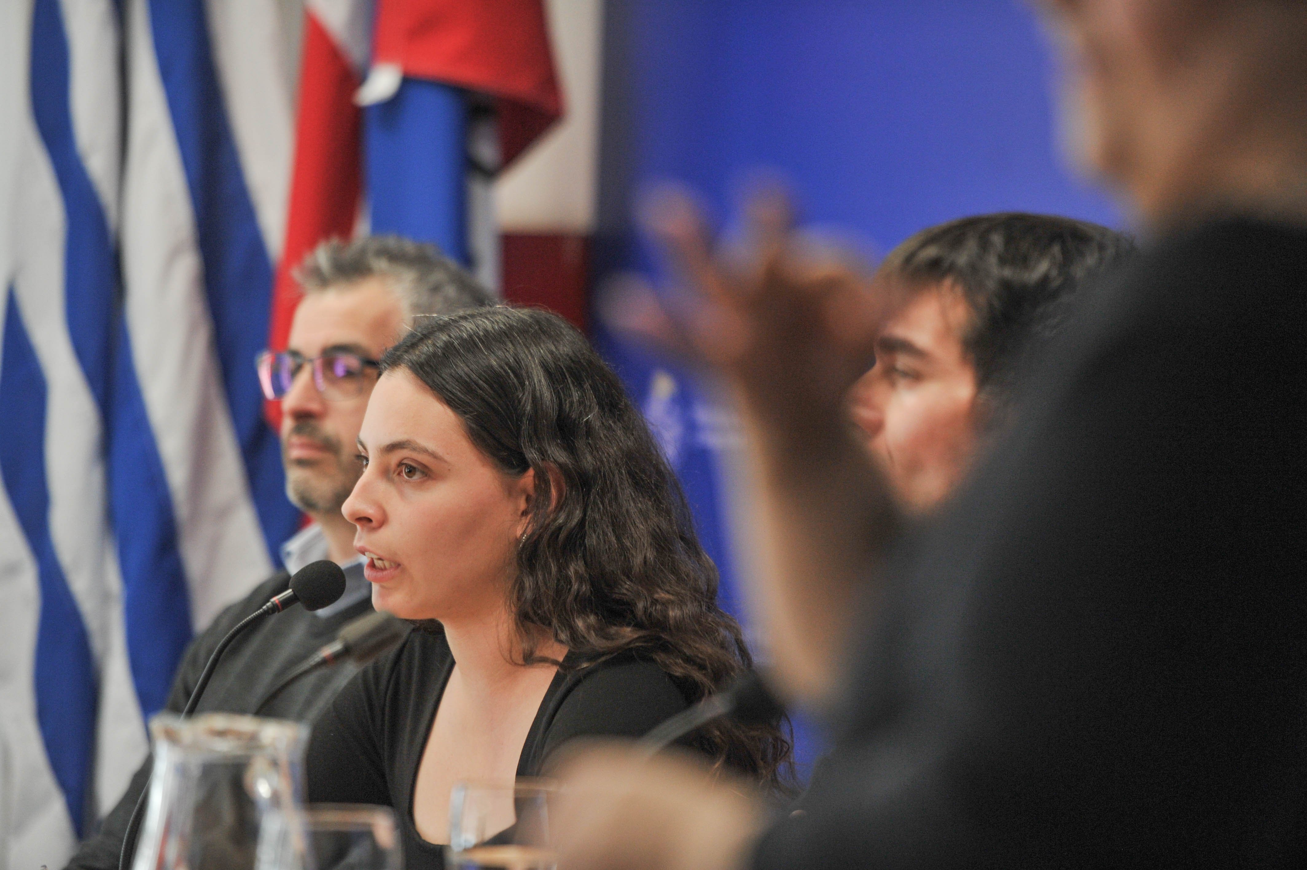 Directora del INJU diserta sentada en mesa con micrófono junto a dos autoridades que la observan.