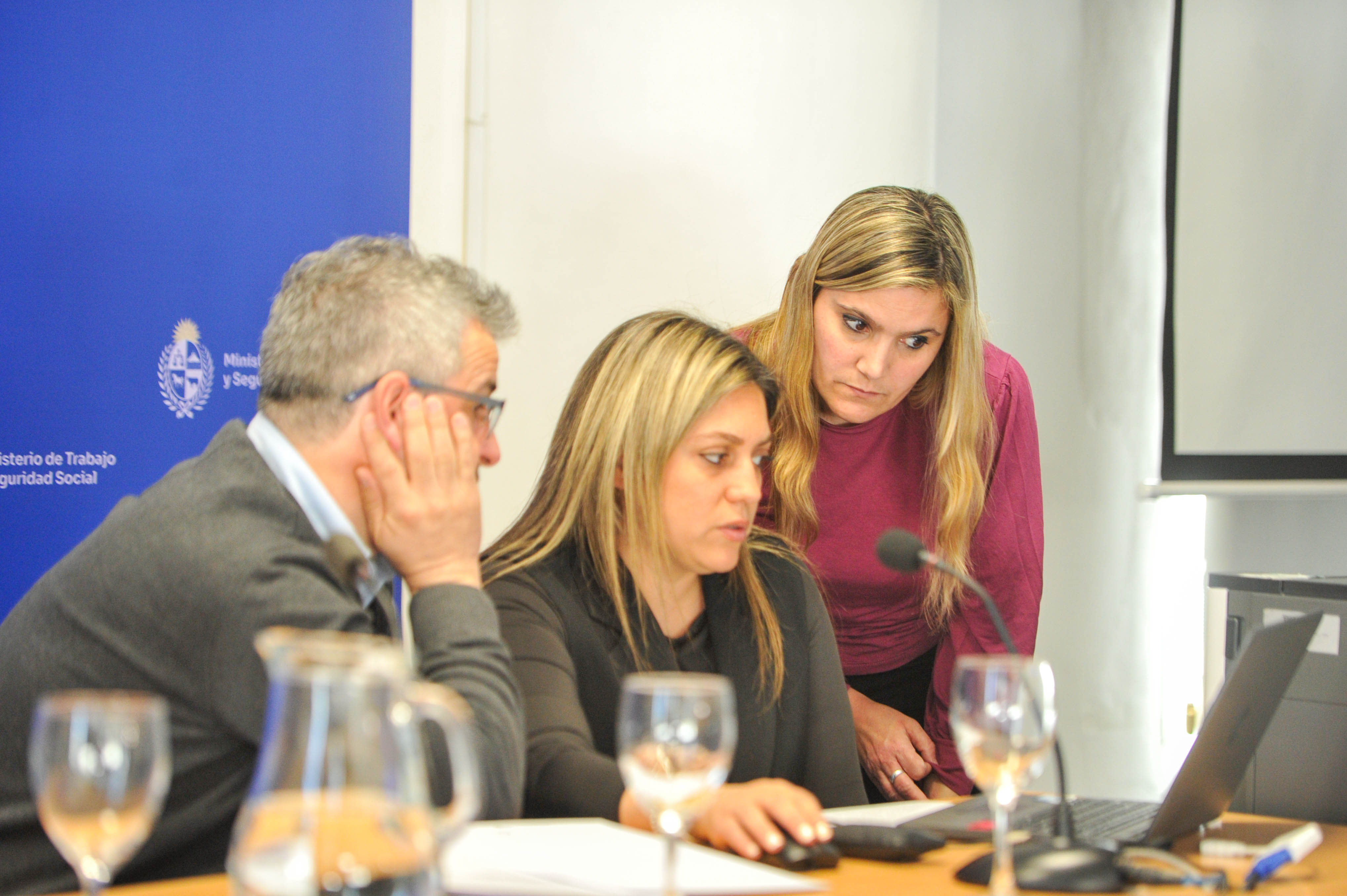 Tres participantes del evento sentados en la mesa principal miran una computadora móvil. Atrás se observa cartel azul con logo del ministerio.