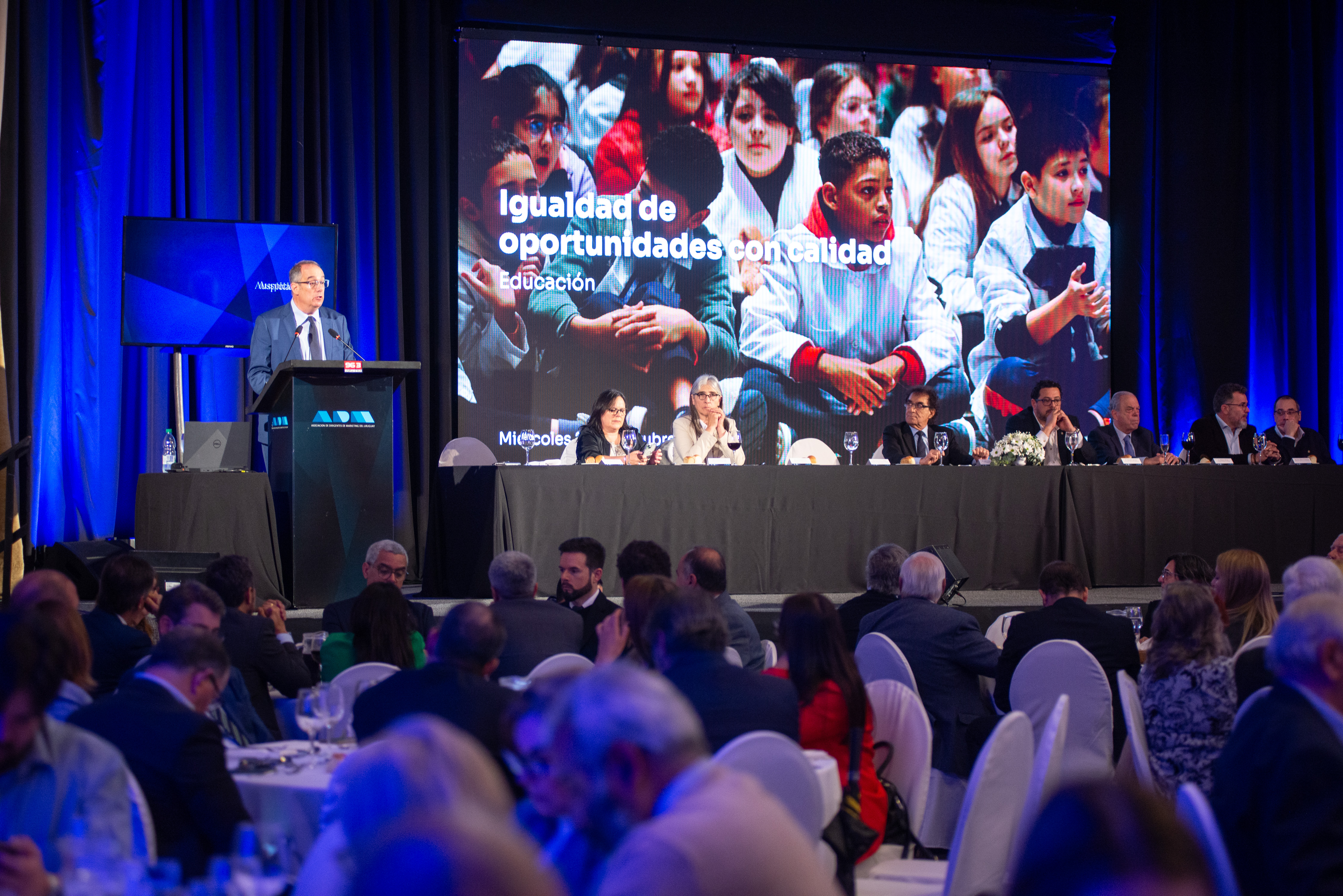 Plano general de la sala de conferencias con el público sentado en mesas circulares mirando al ministro de Educación que diserta de pie en atril.