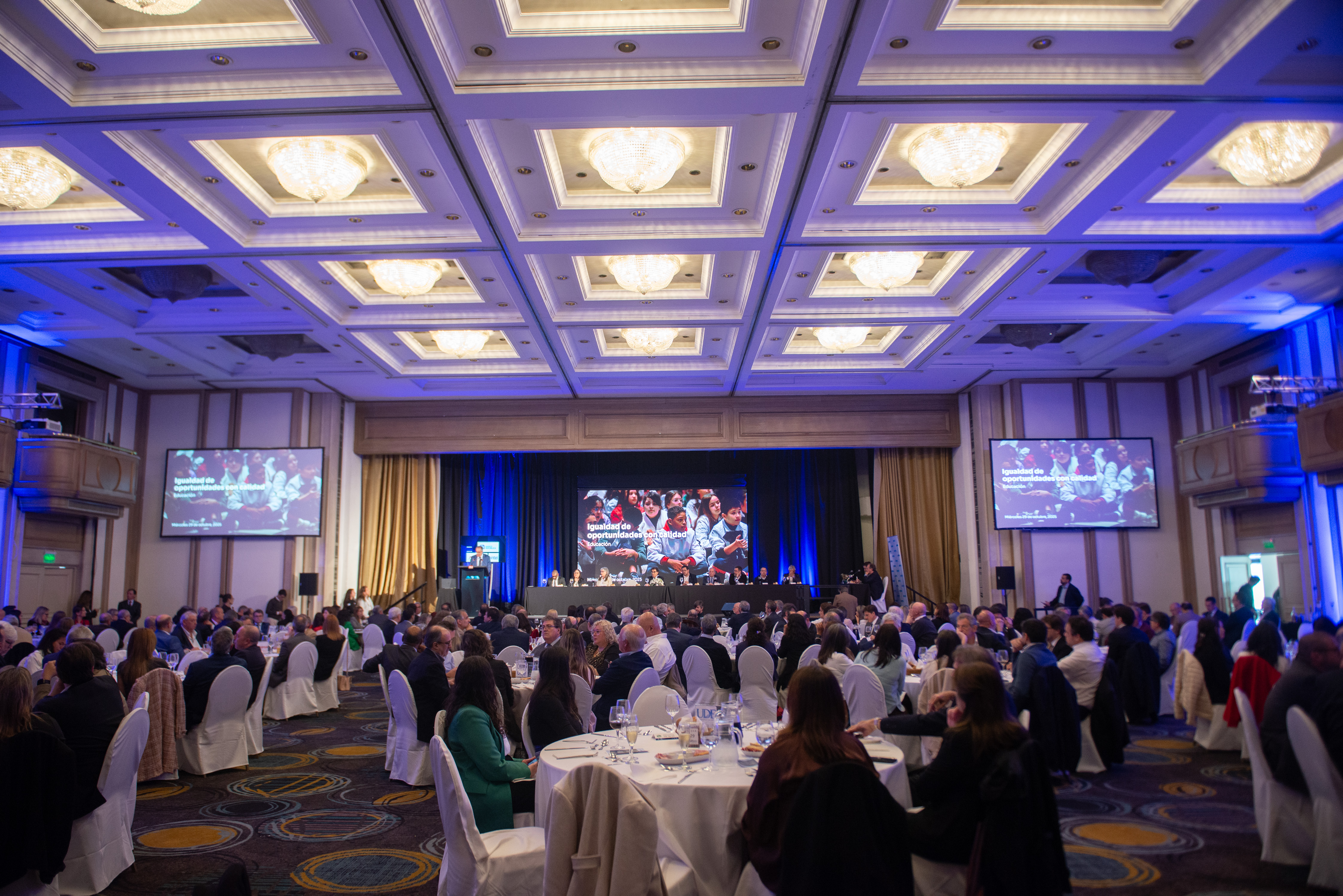 Plano general de la sala de conferencias con el público sentado en mesas circulares mirando al ministro de Educación que diserta de pie en atril.