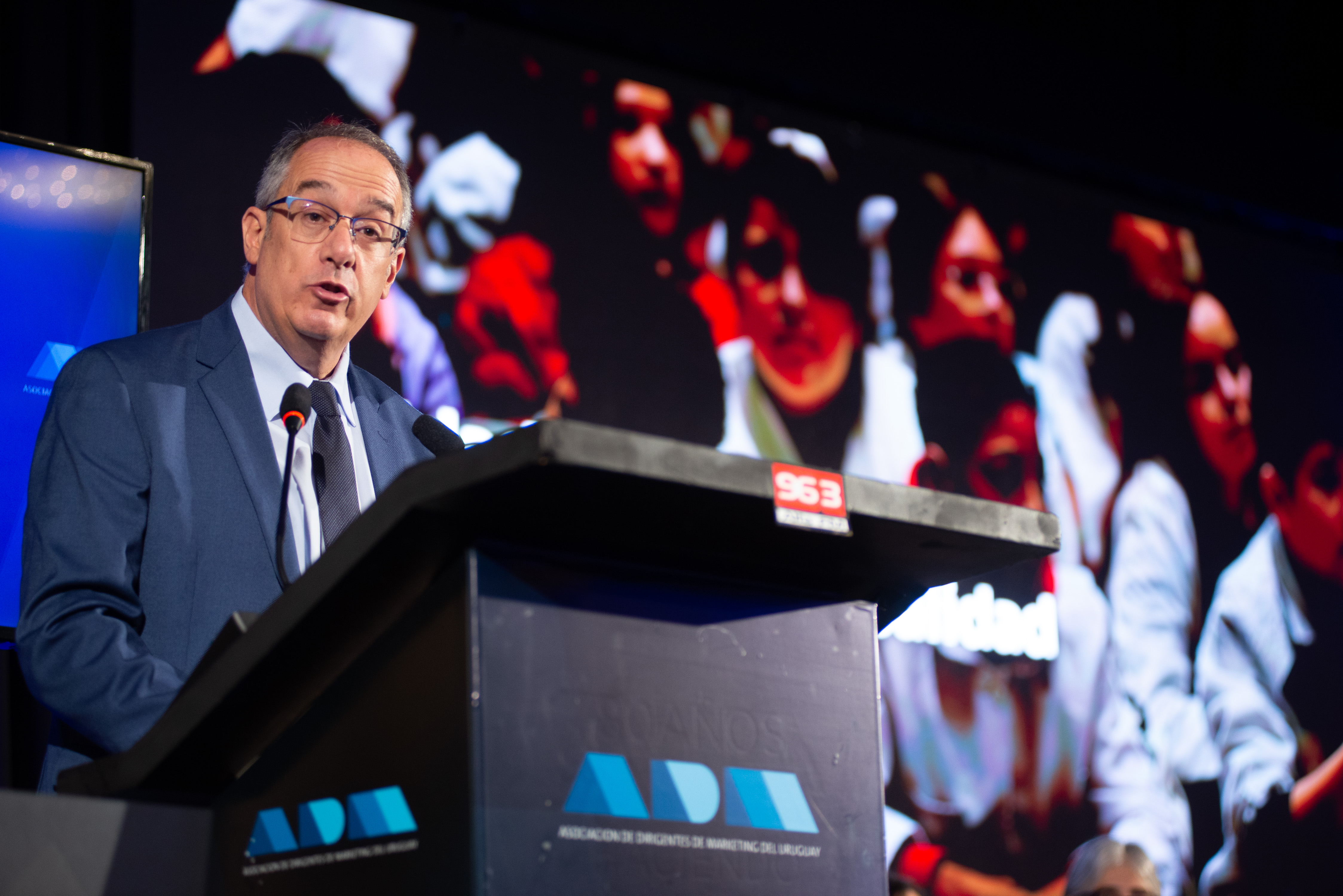 Ministro de Educación diserta de pie en atril con micrófono en el escenario. Atrás se observa gran pantalla con imagen de varios escolares.