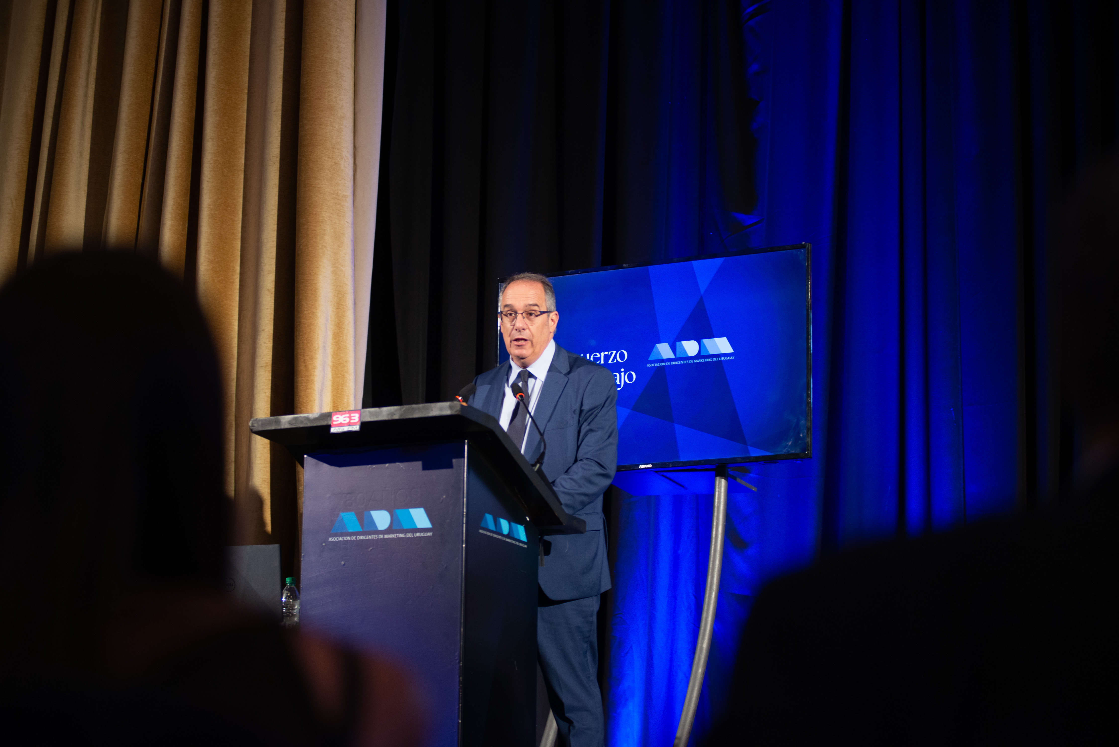 Otra perspectiva del ministro de Educación disertando de pie en atril con micrófono. Atrás se observa telón azul y pantalla que dice ADM.