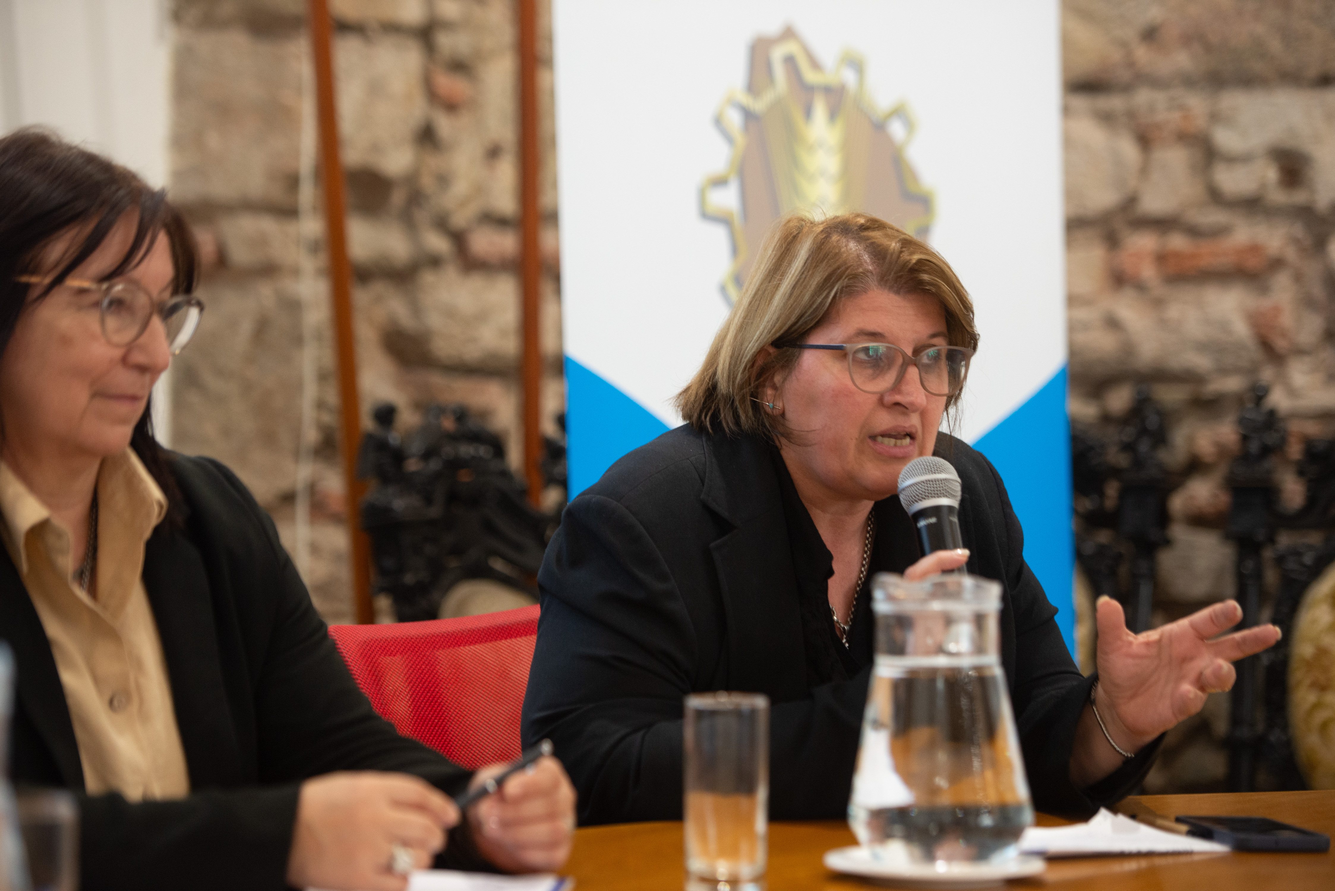 Directora general de UTU diserta sentada en mesa con micrófono. La consejera de la ANEP la escucha atentamente a su lado.