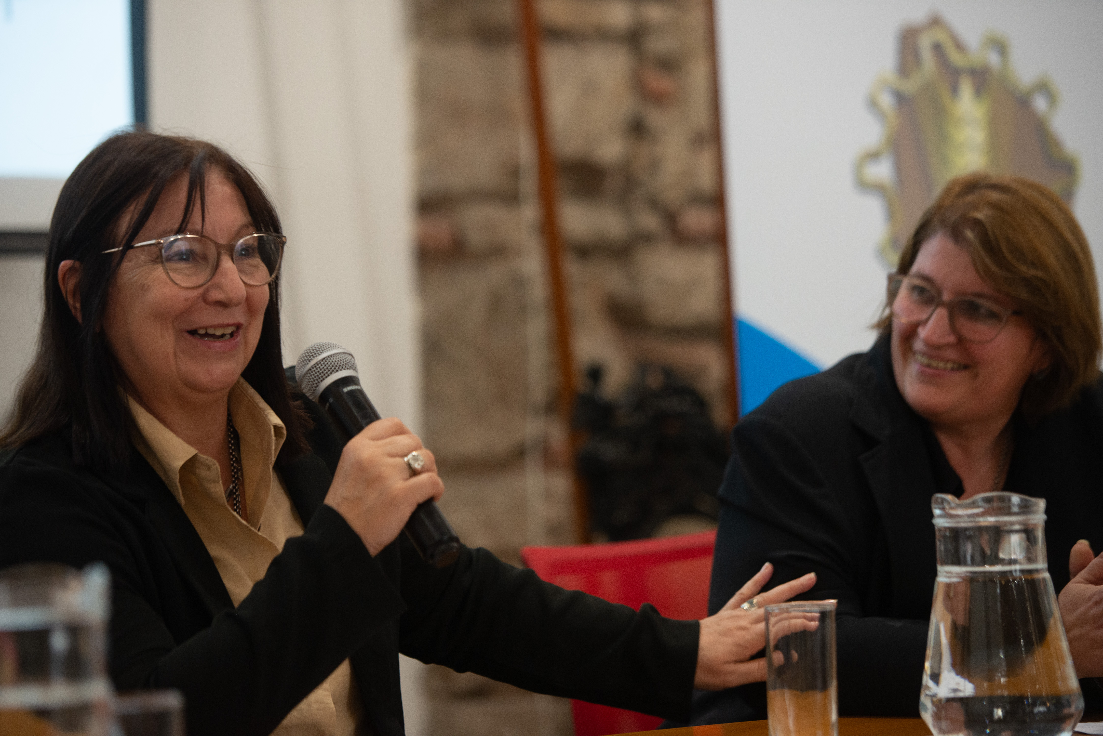 La consejera de la ANEP diserta sentada en mesa con micrófono mientras la directora general de UTU la observa, sonriente.