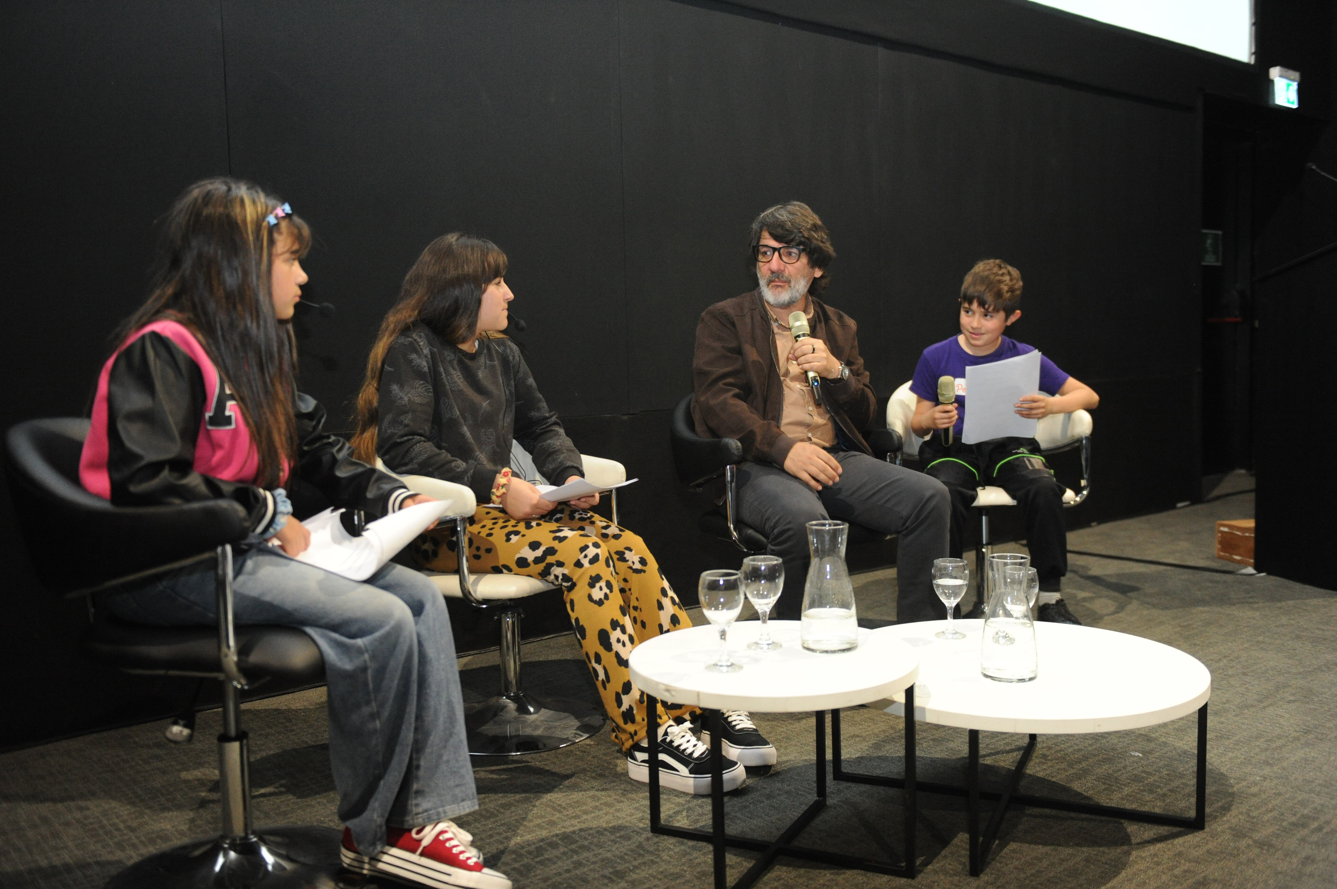 El actor Cesar Troncoso dialoga con tres niños en el escenario sentados en sillones con una mesa pequeña en el centro con vasos de agua.