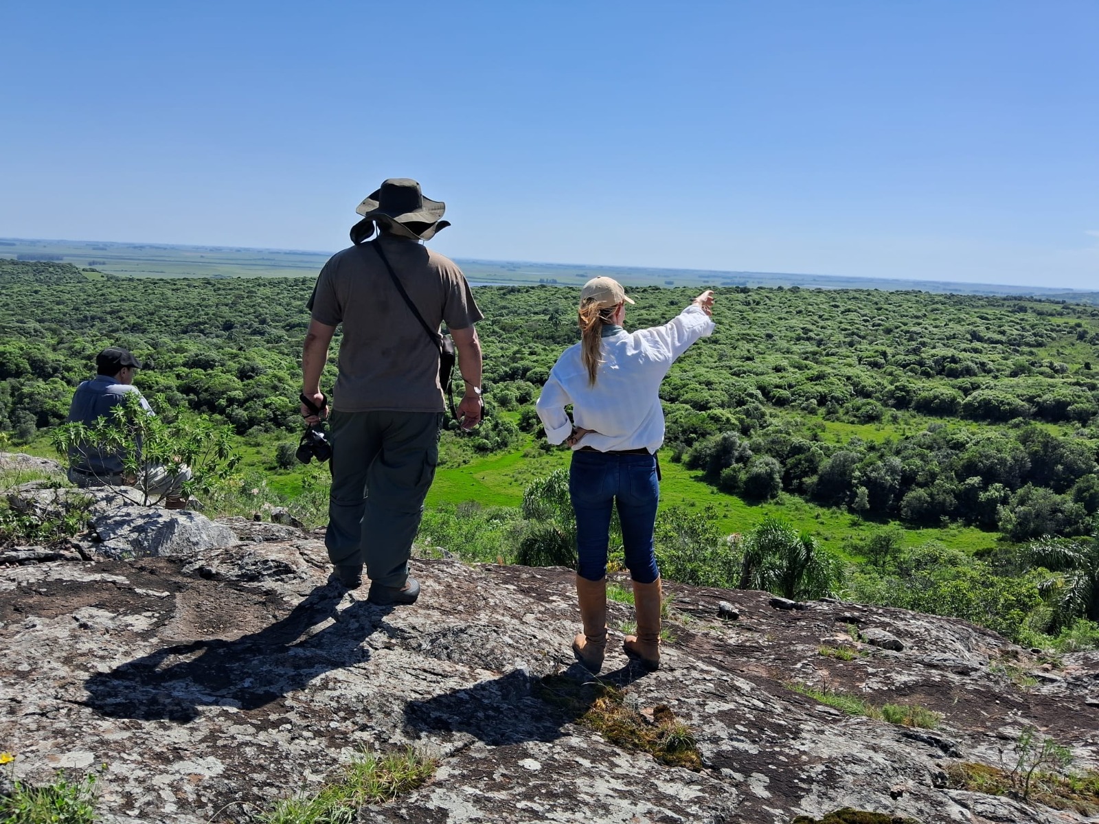 Una mujer y un hombre en lo alto de una sierra observan la inmensidad del monte nativo y el horizonte, bajo un cielo azul.