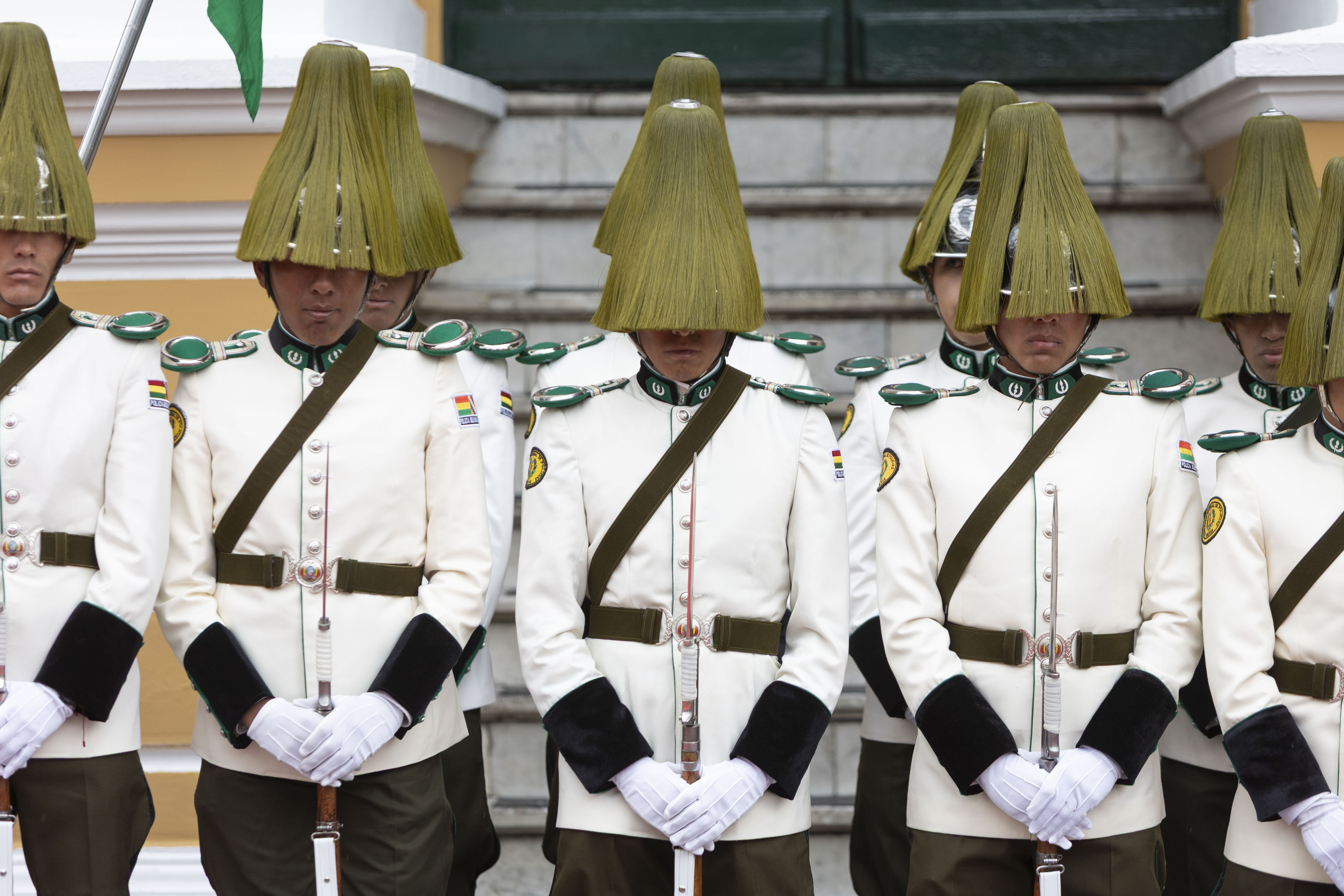 Guardias de honor bolivianos, en formación con uniformes blancos y cascos de flecos de palma verde que cubren sus rostros.