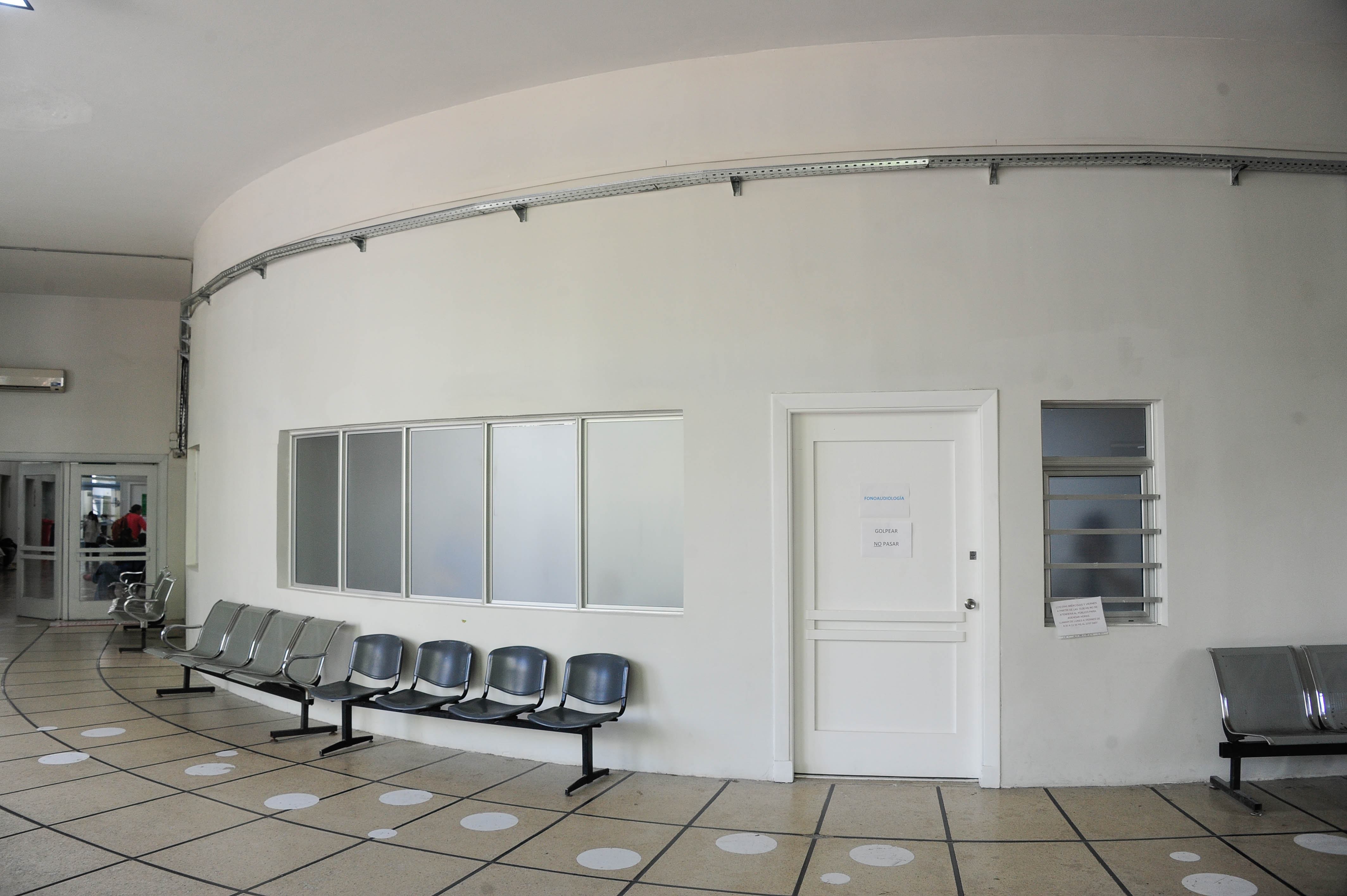 Plano general de una sala de espera con fila de sillas grises apoyadas en paredes blancas.