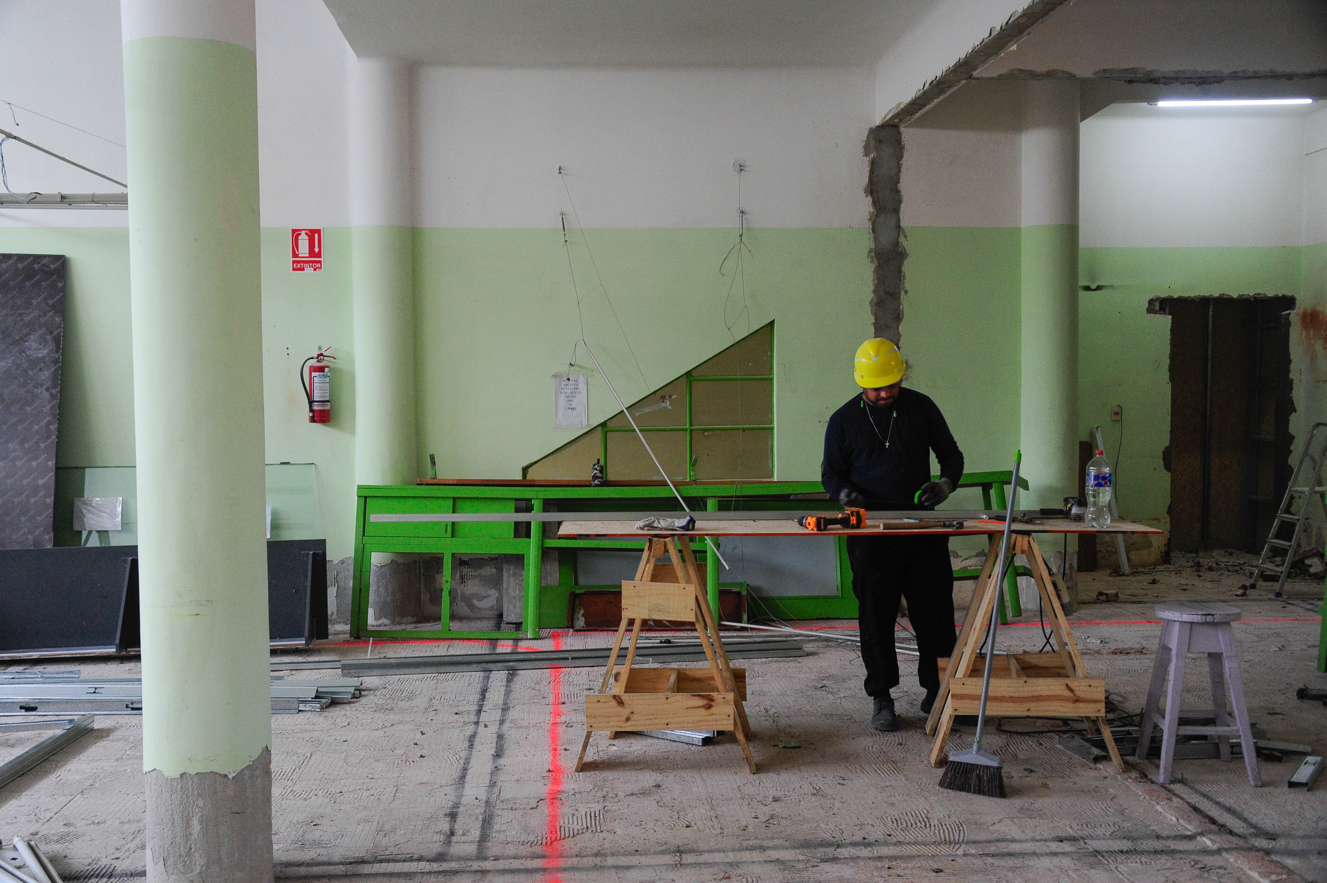 Un obrero con casco amarillo trabaja en una mesa de caballetes en un amplio espacio interior en remodelación, con paredes verdes y blancas.