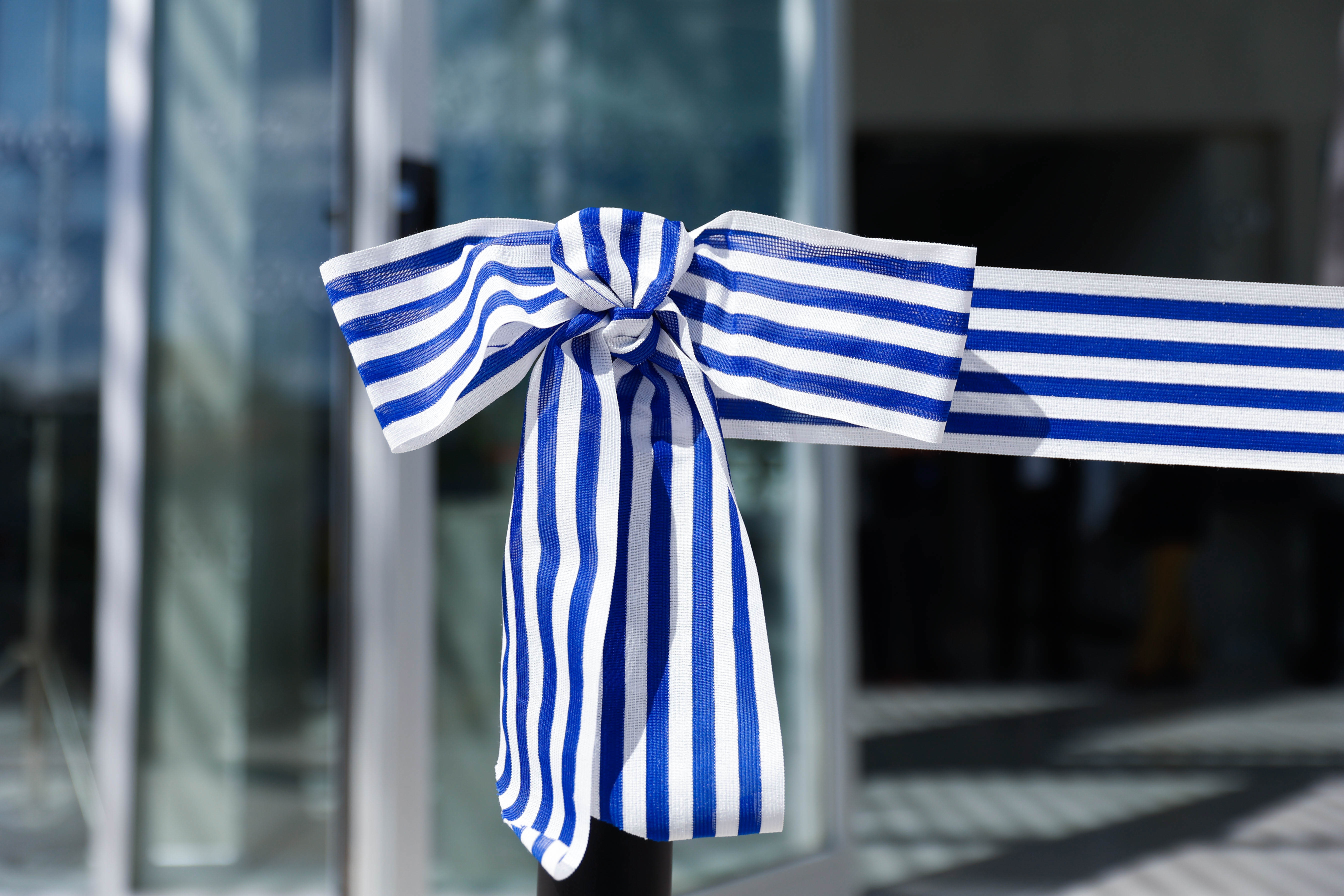 Primer plano de una moña blanca y azul a rayas, atada a una cinta de inauguración que bloquea la entrada del edificio de la UTEC.