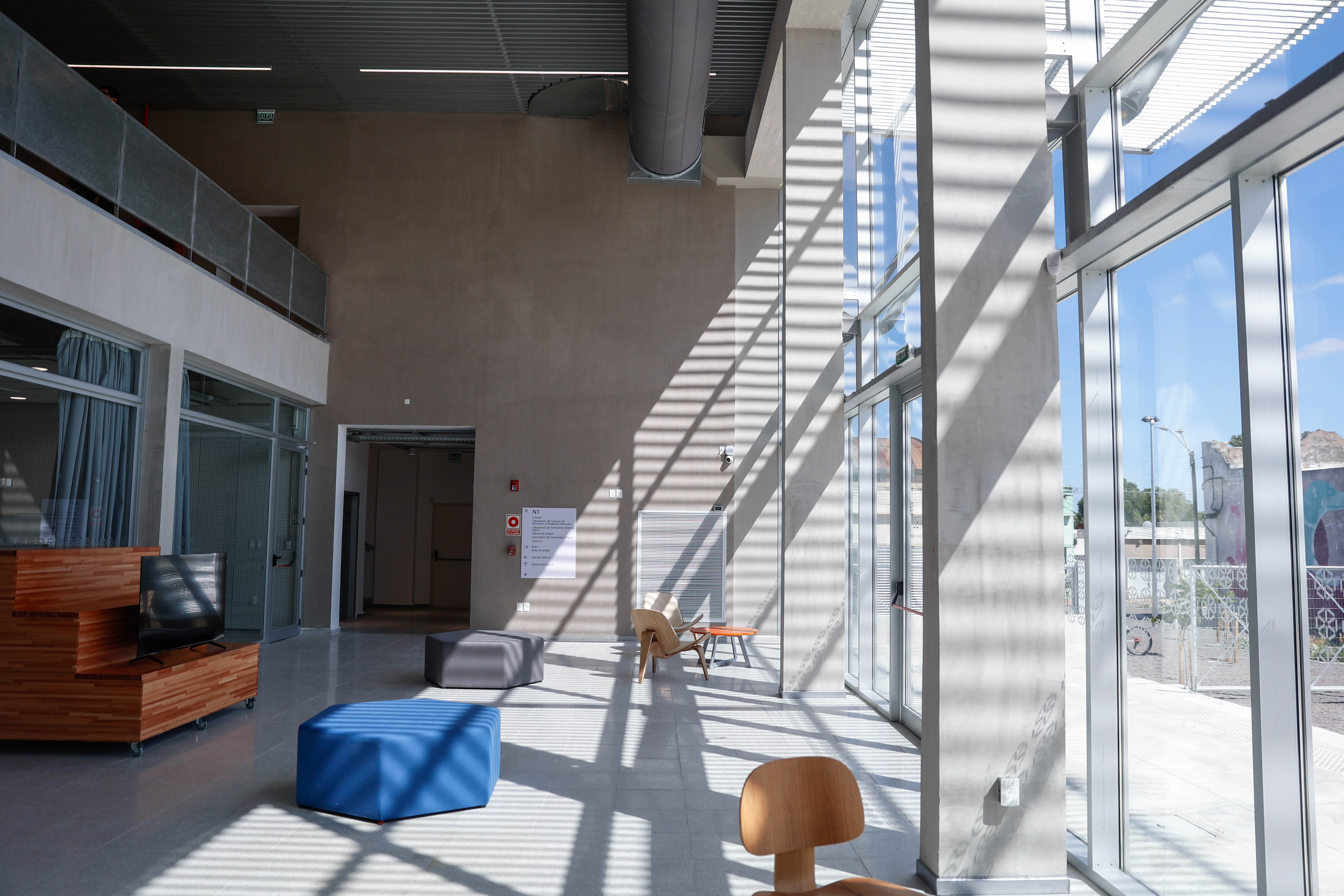 Sala de ingreso del nuevo edificio de la UTEC en Lavalleja con ventanales amplios, sillas de diseño y un mueble de madera modular con televisor.