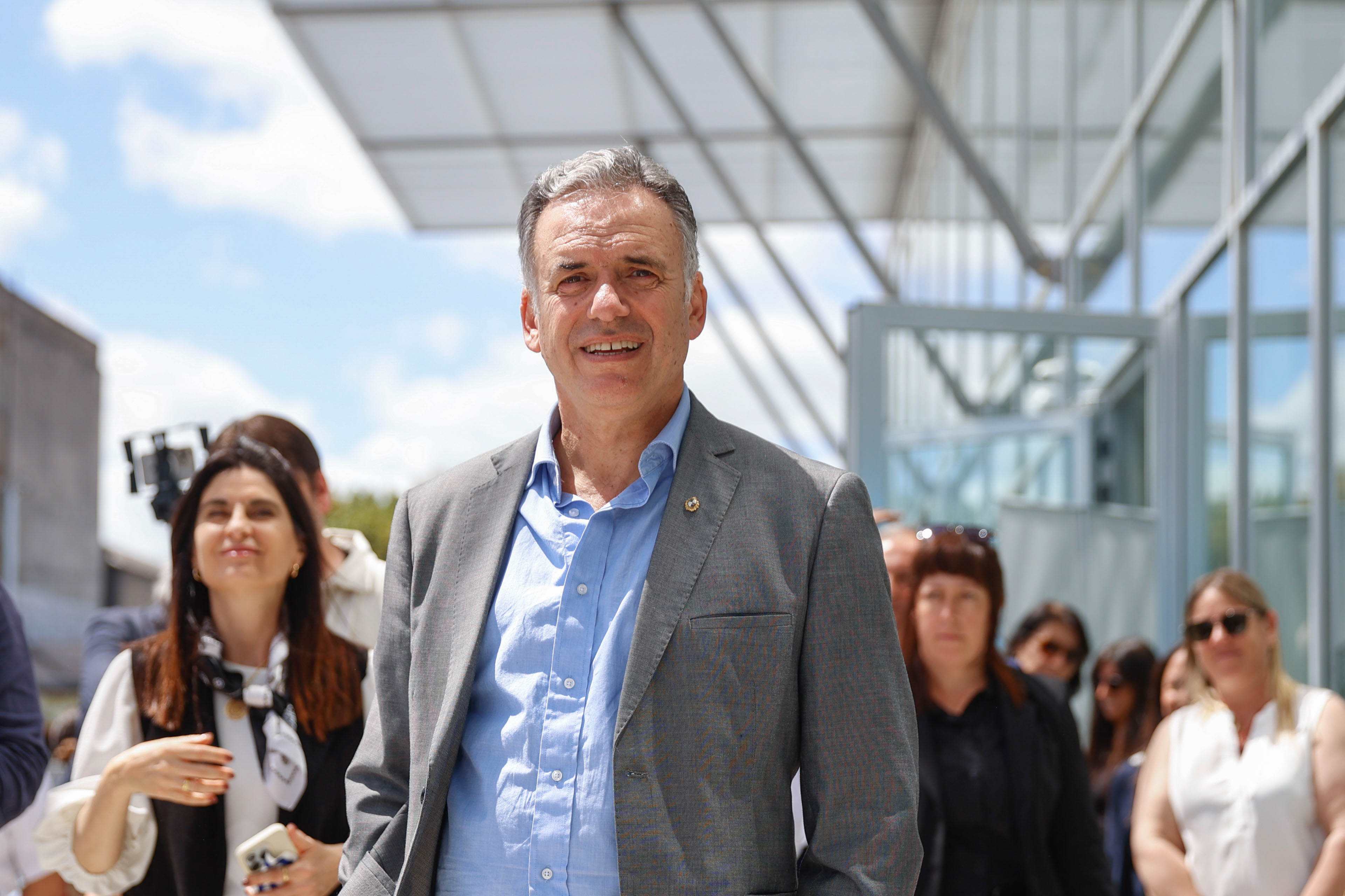 Orsi, sonriente y vestido con saco gris, posa frente a cámara en el evento al aire libre rodeado de gente.