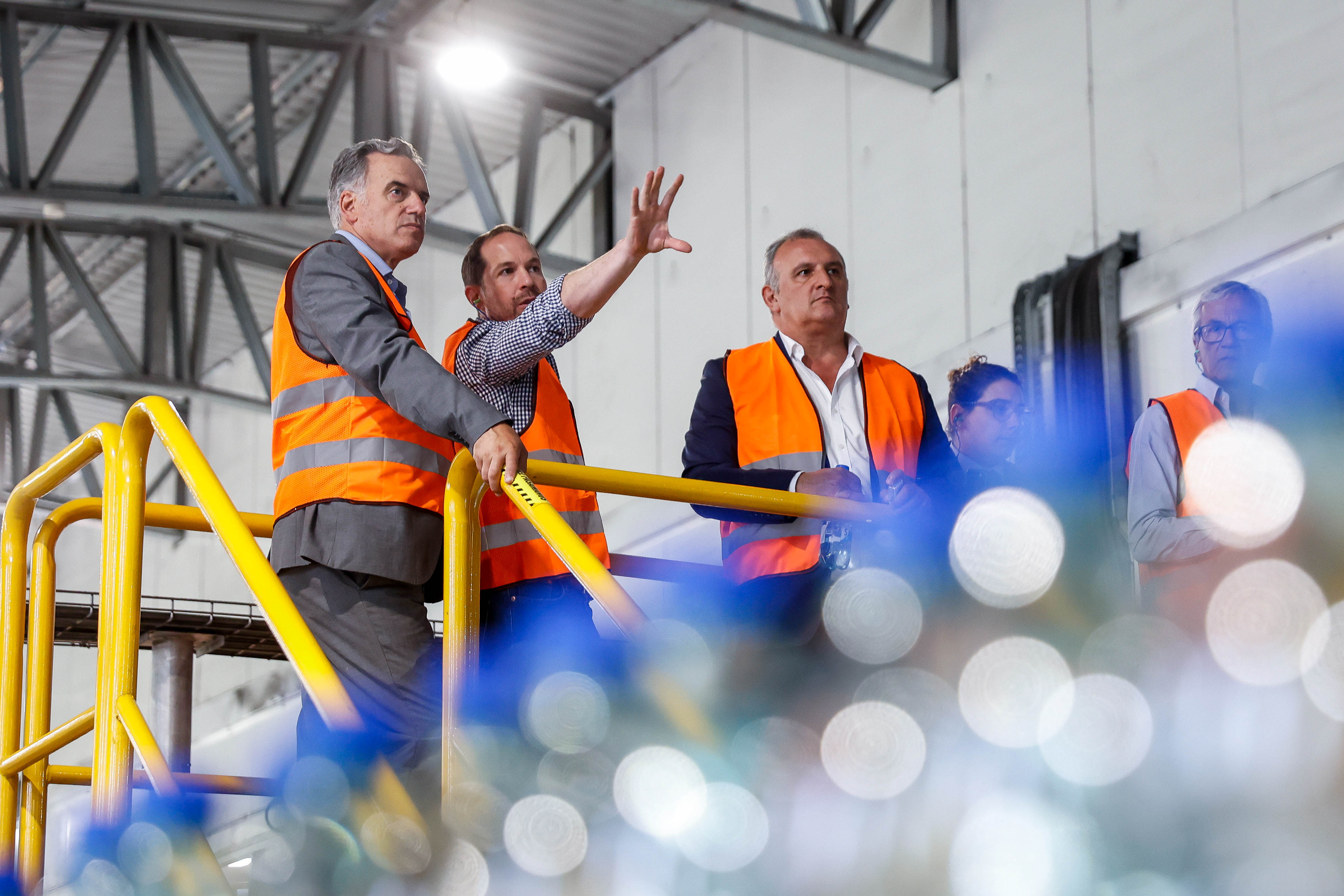Orsi y dos hombres con chalecos naranjas de seguridad observan y discuten procesos industriales desde una pasarela elevada en una planta.