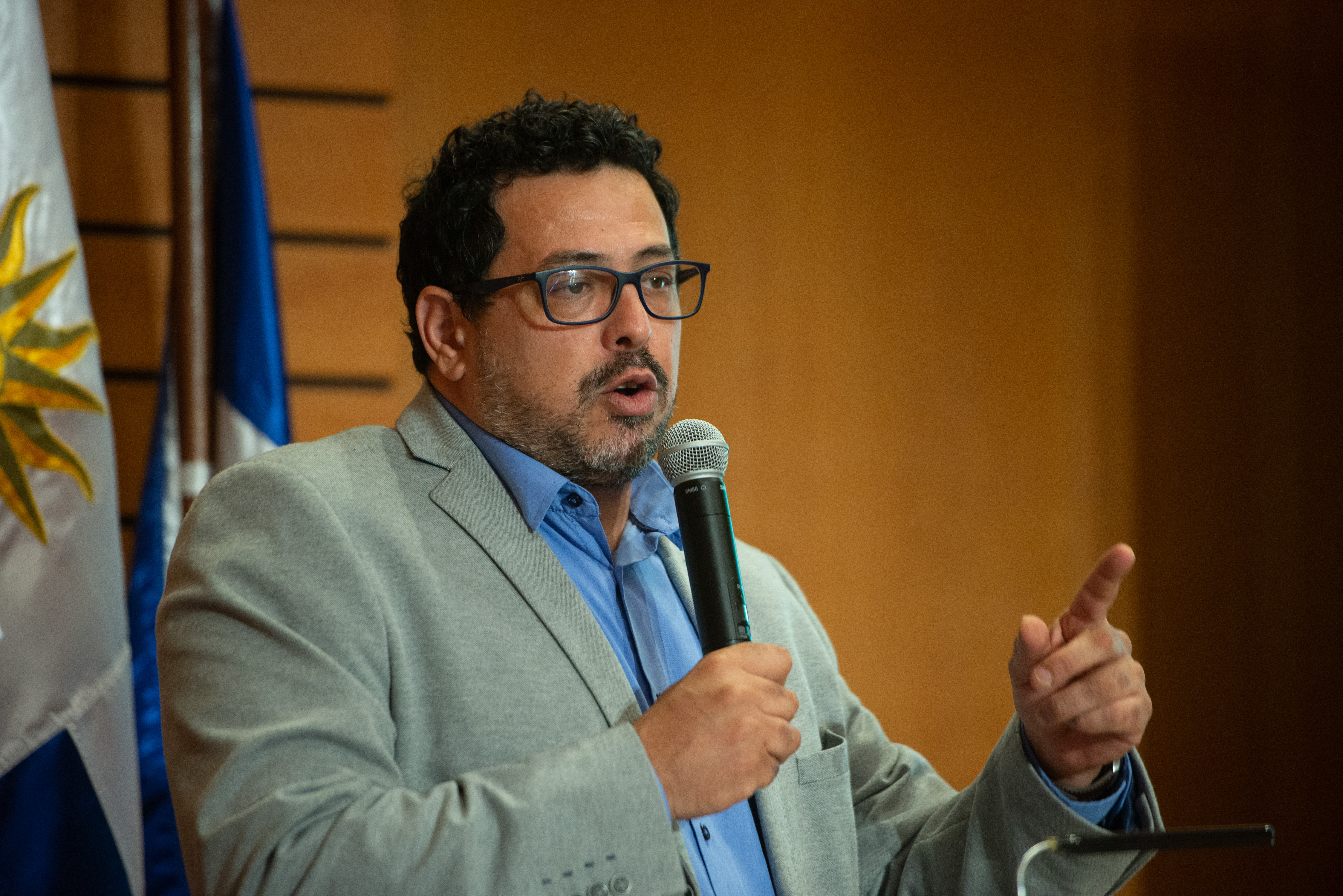 Secretario de Presidencia diserta de pie en atril con micrófono en mano. Atrás se observa el pabellón nacional.