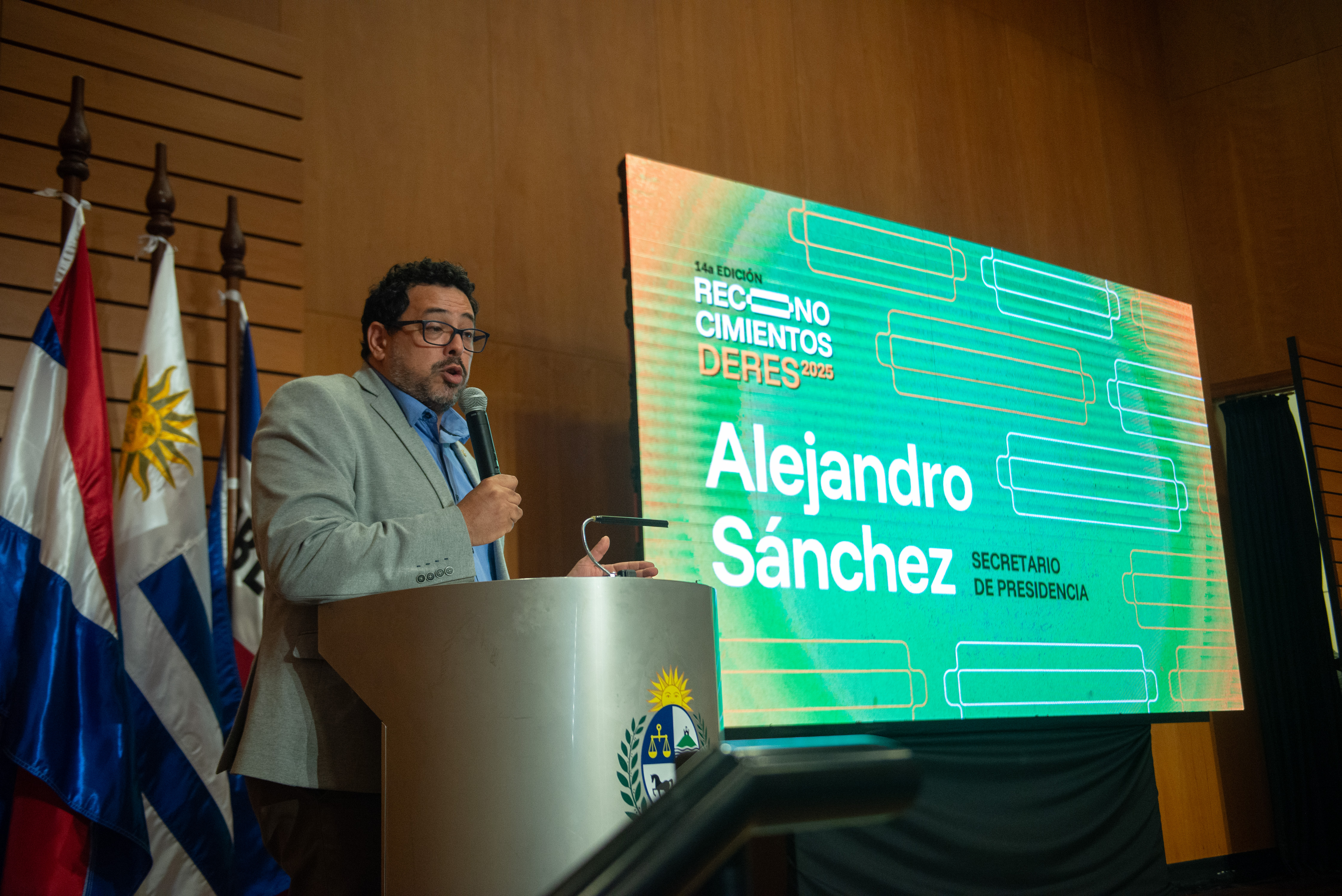 Secretario de Presidencia diserta de pie en atril con micrófono en mano. Atrás se observan los pabellones patrios y pantalla con el nombre de él.