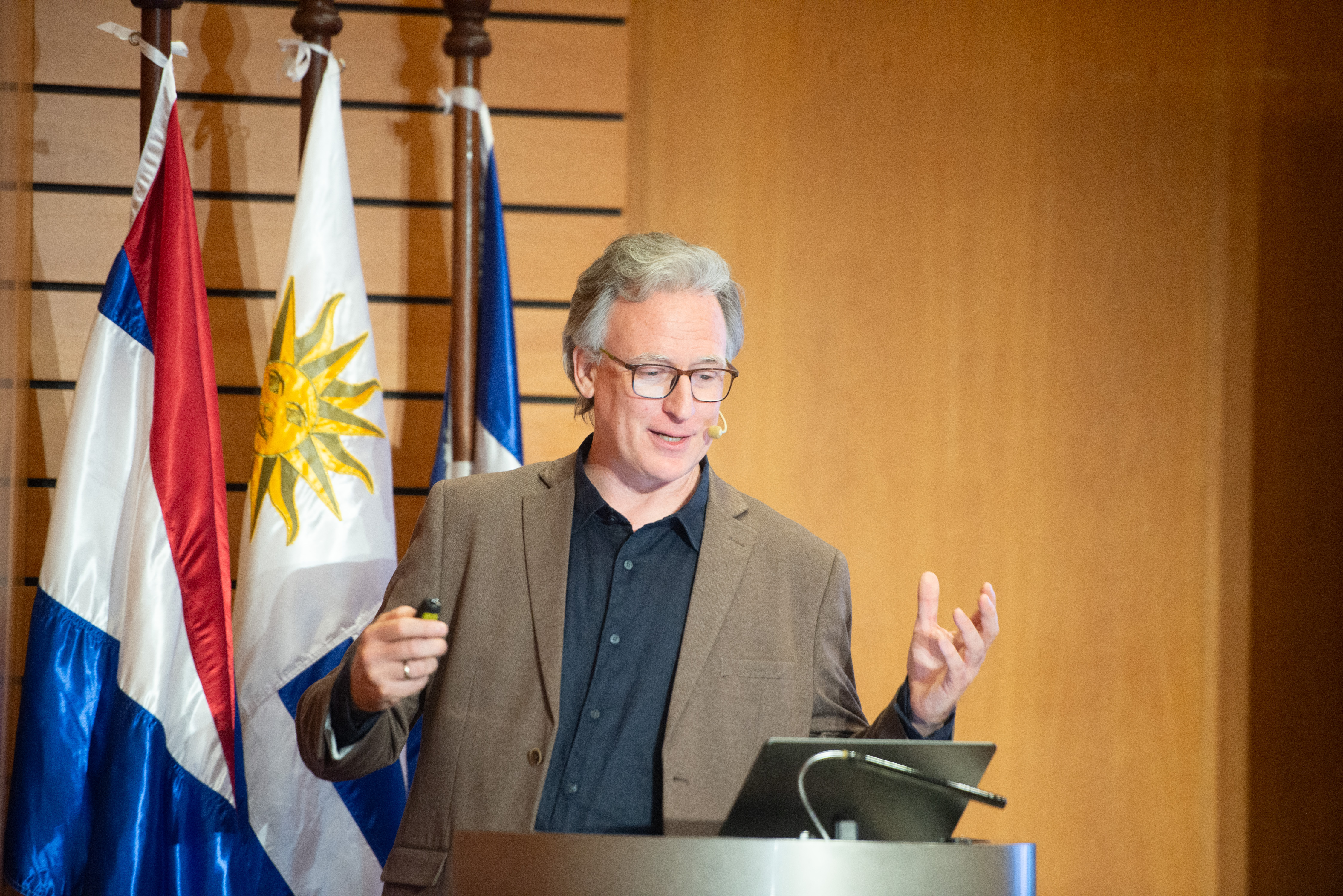 Representante de Deres diserta de pie en atril con micrófono. Atrás se observan los pabellones patrios.