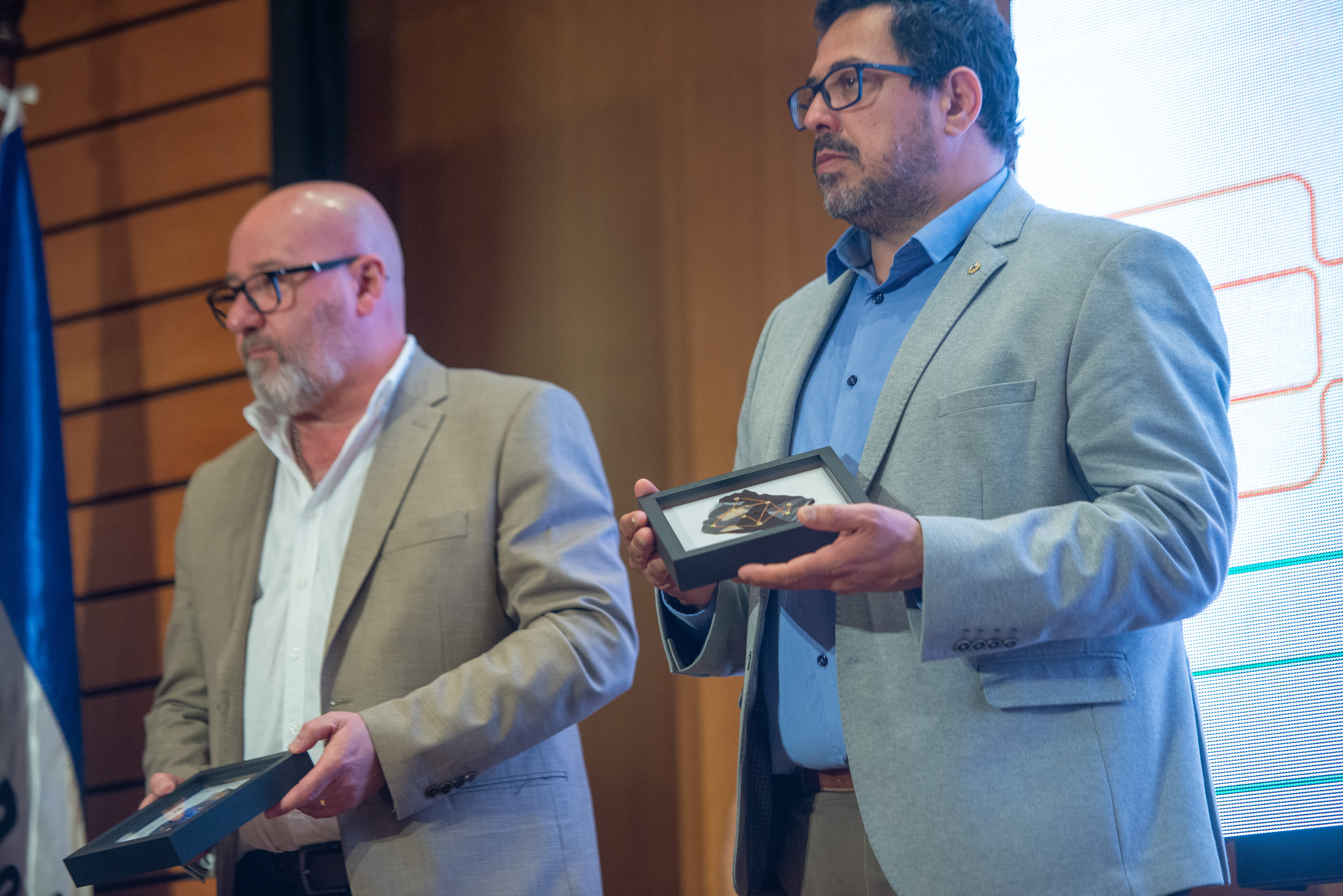 Secretario de Presidencia junto a un representante de Deres de pie en el escenario sosteniendo un cuadro de obsequio.