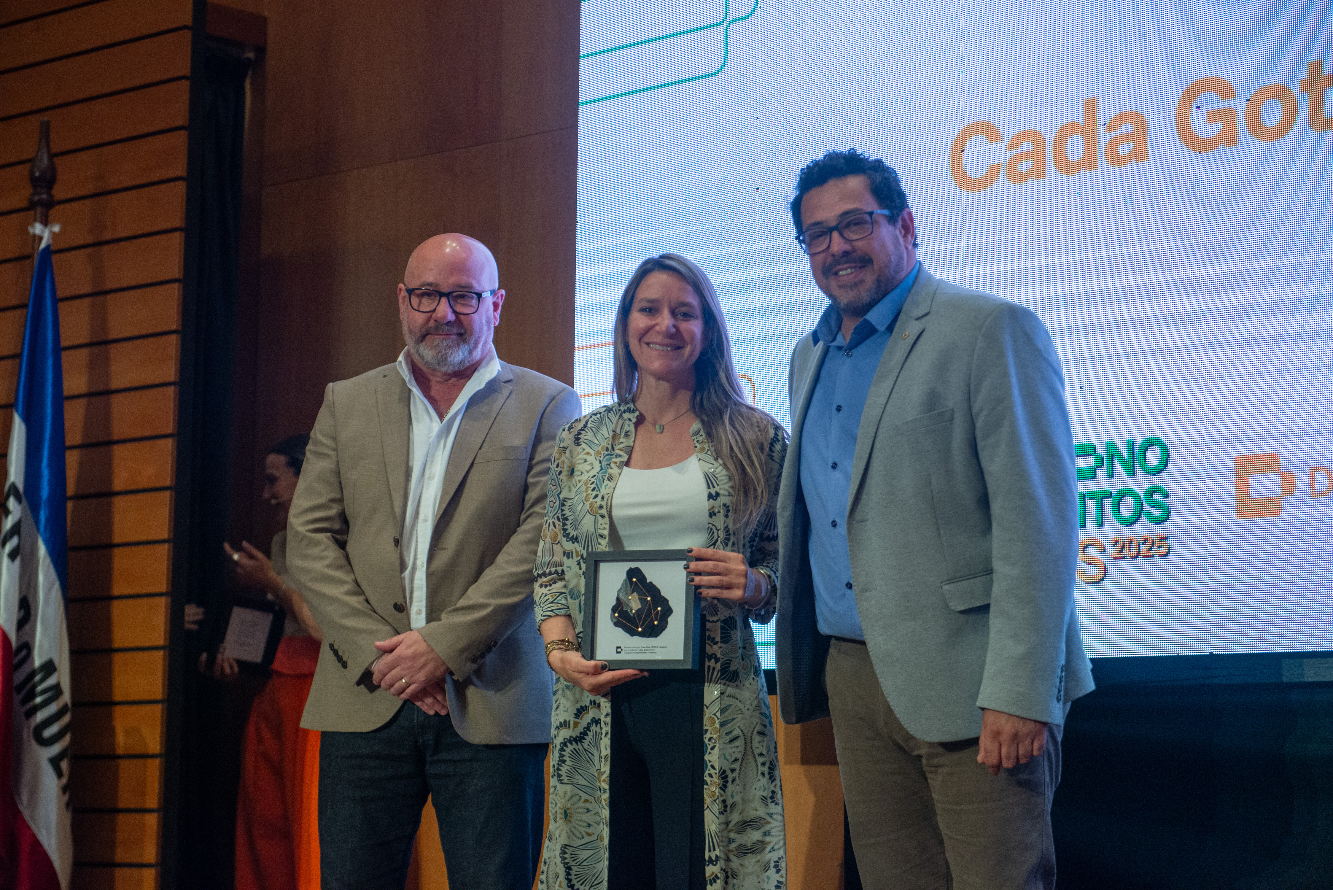 Secretario de Presidencia junto a dos representantes de Deres de pie en el escenario, uno de ellos sostiene cuadro.