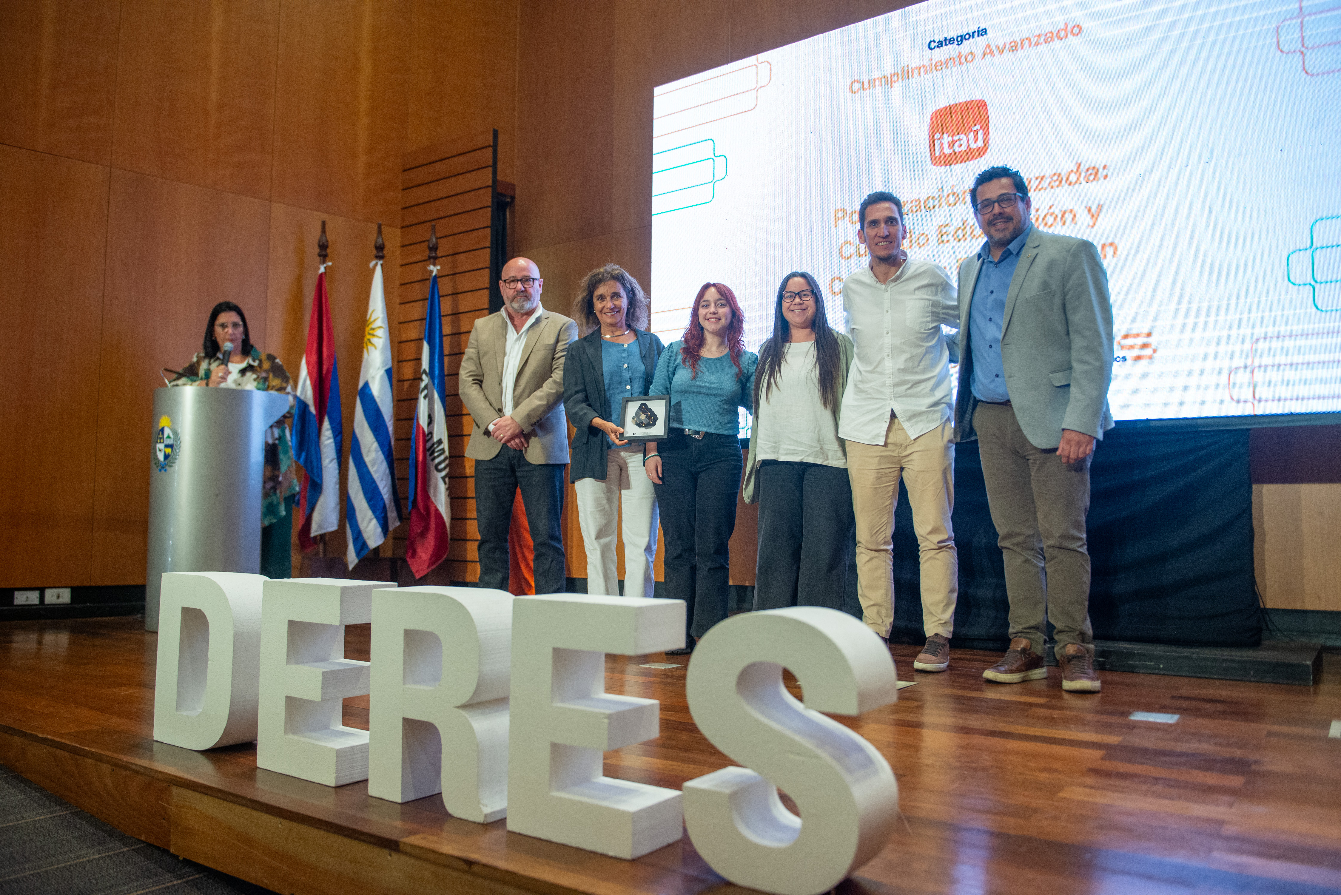 Secretario de Presidencia, junto a representantes de Deres, de pie en el escenario posa sonriente frente a cámara.