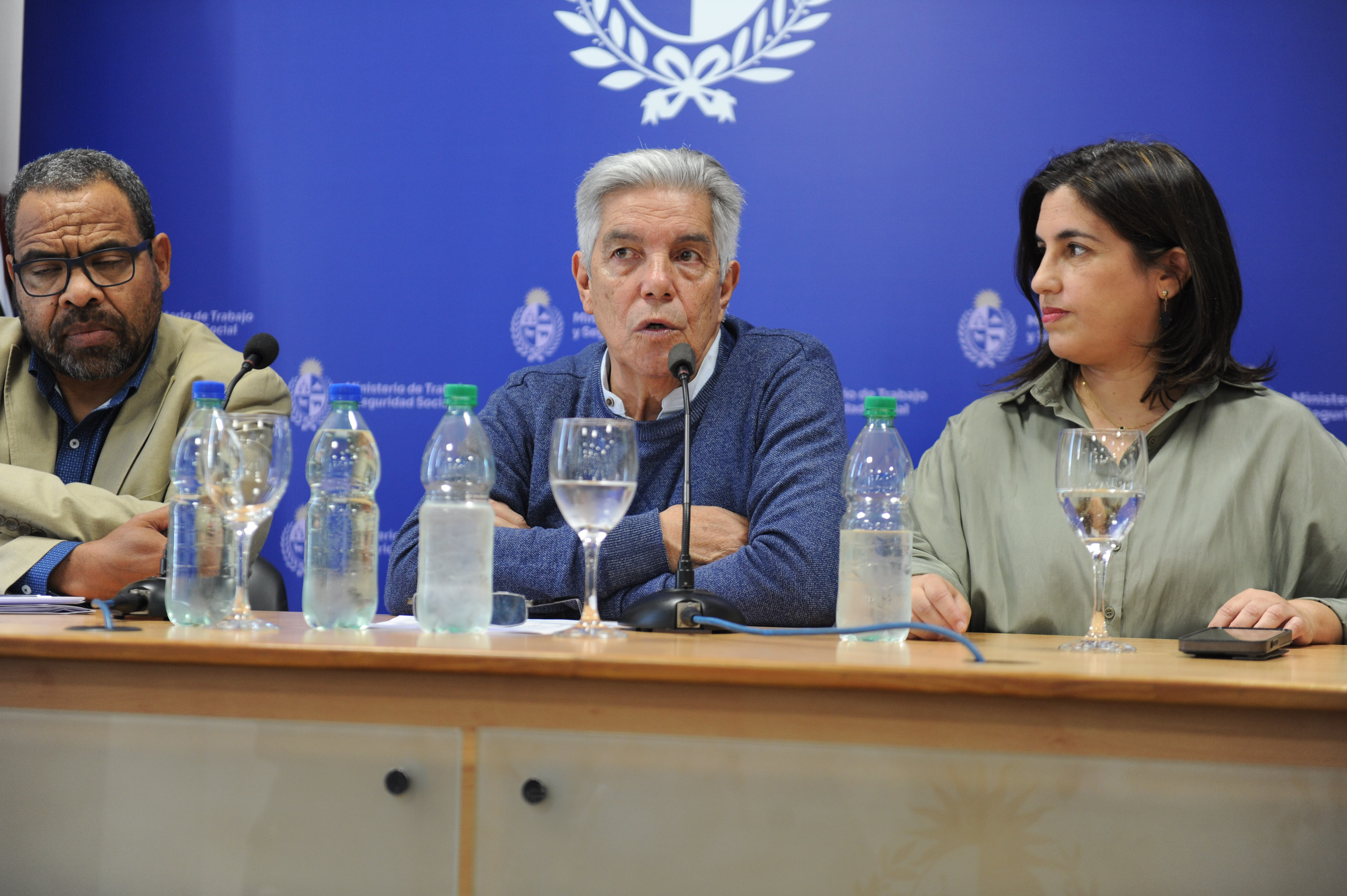 Inspector general de Trabajo y Seguridad Social diserta sentado en mesa con micrófono. A sus costados, dos autoridades lo observan.
