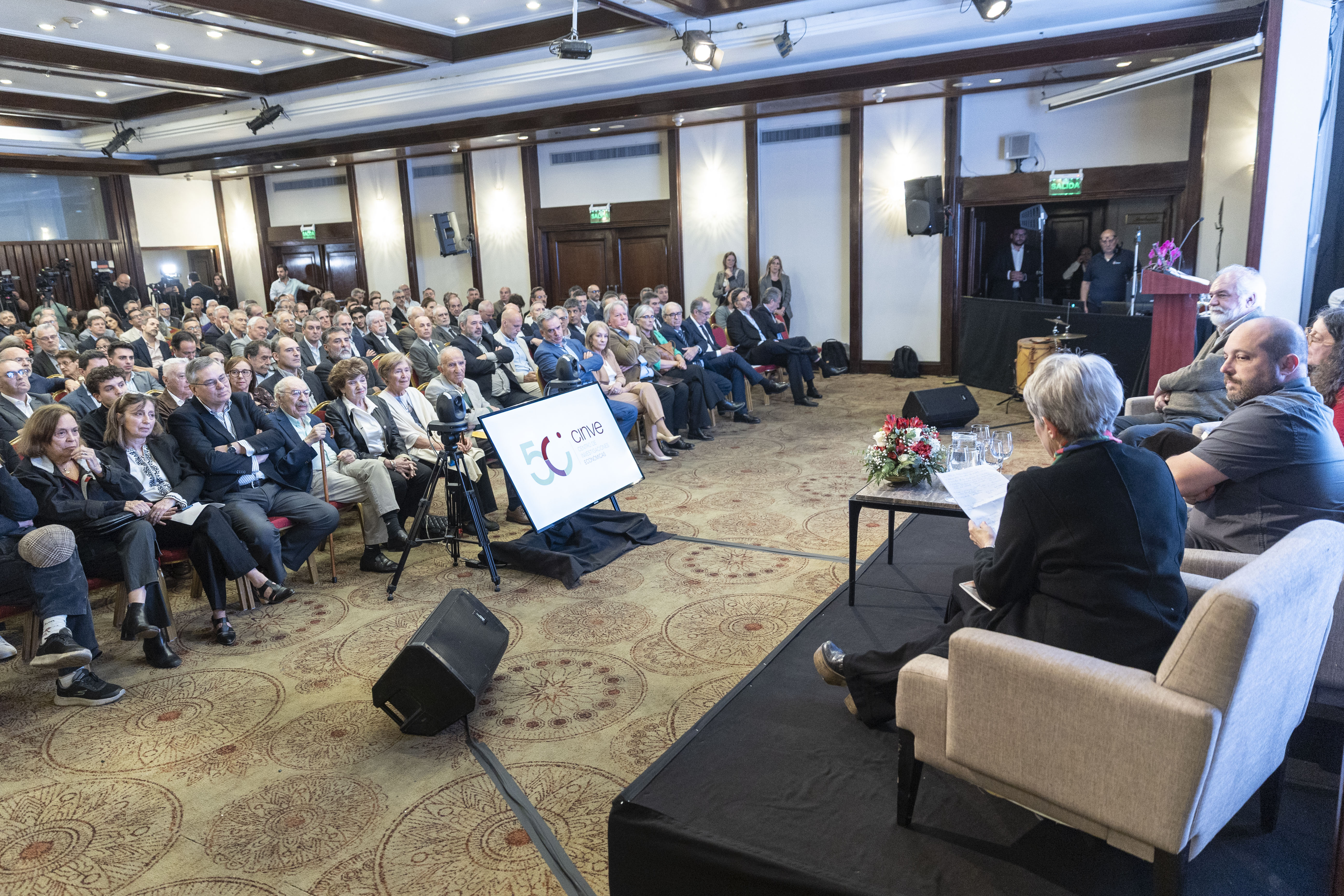 Plano general de sala de reuniones con el público sentado en la platea mirando hacia el escenario donde las autoridades disertan.