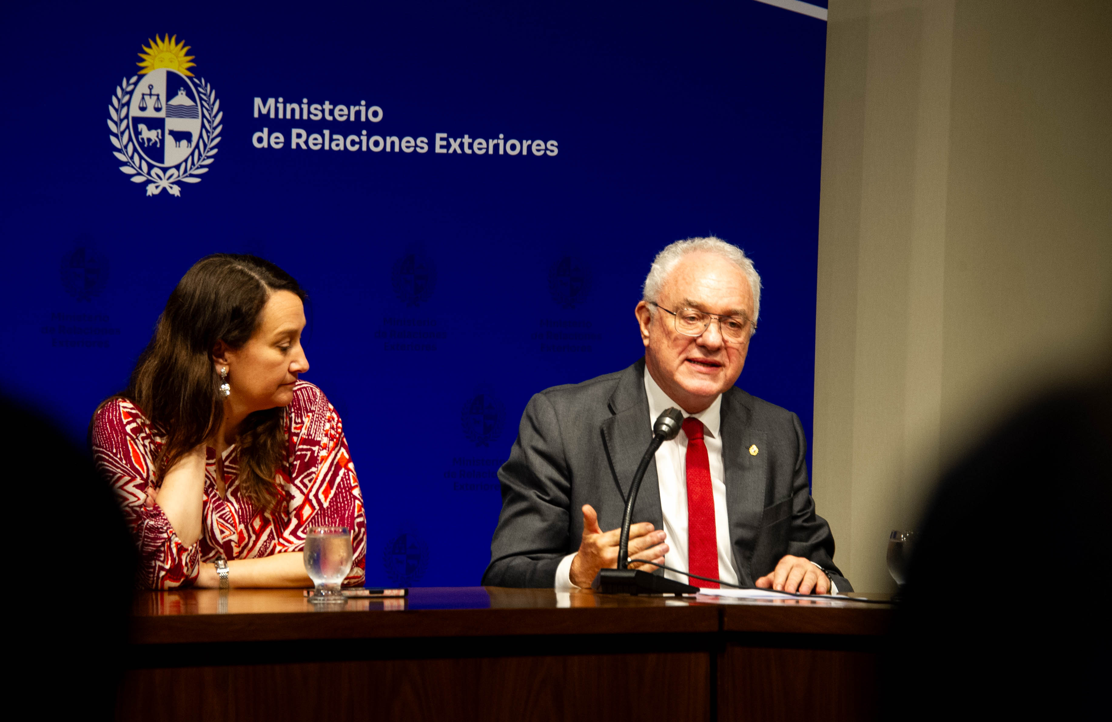 Canciller diserta sentado en mesa con micrófono, a su lado está la vicecanciller que lo mira atentamente.