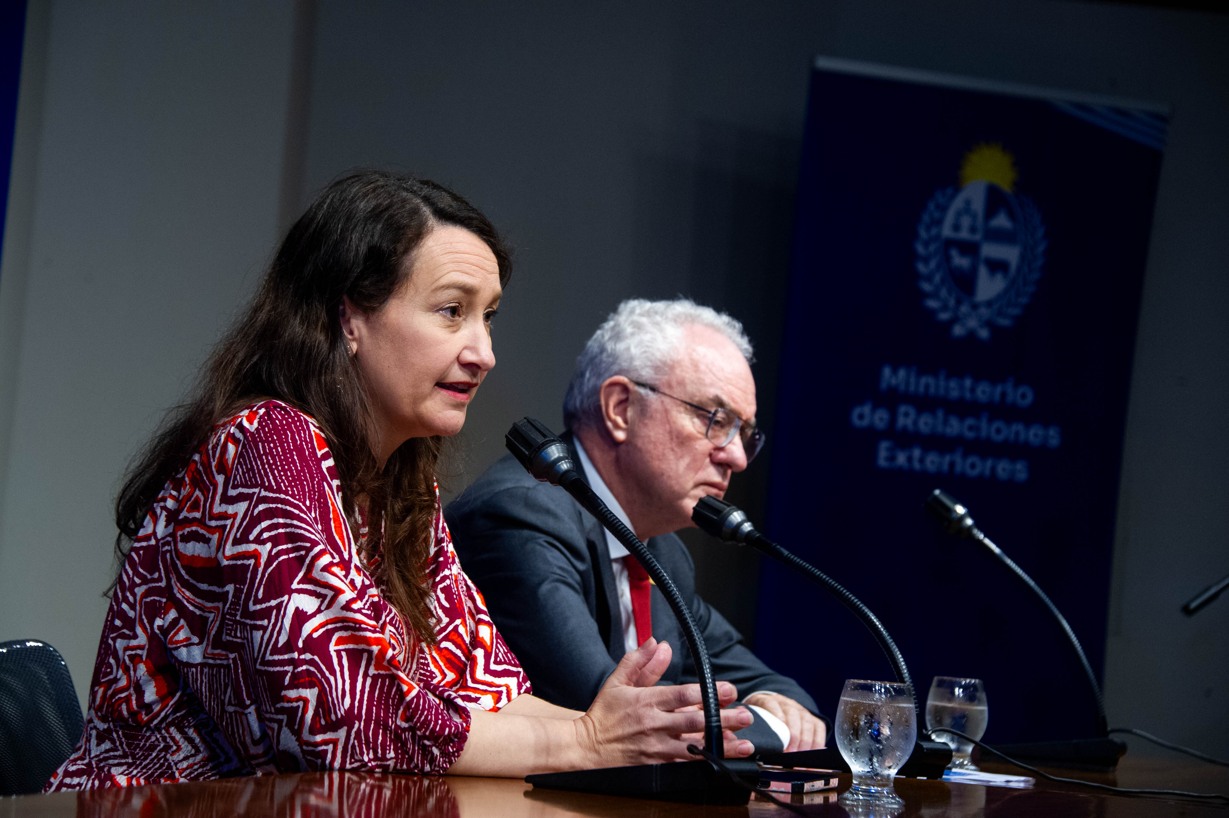 La vicecanciller, de perfil, diserta sentada en mesa con micrófono. A su lado está el canciller quien escucha atentamente.
