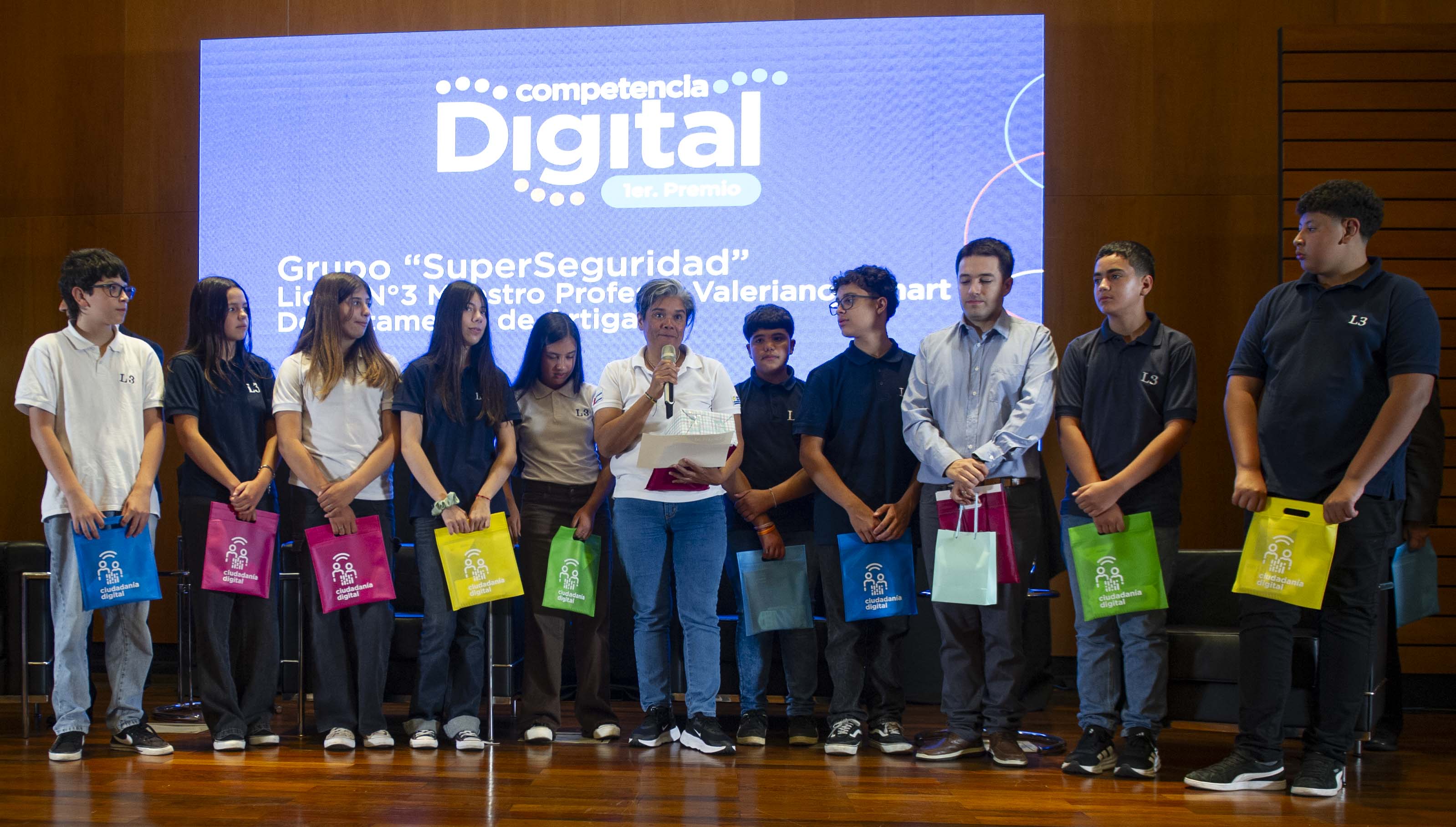 Docente diserta de pie en el escenario con micrófono en mano junto a un grupo de estudiantes que sostienen bolsas de regalos.