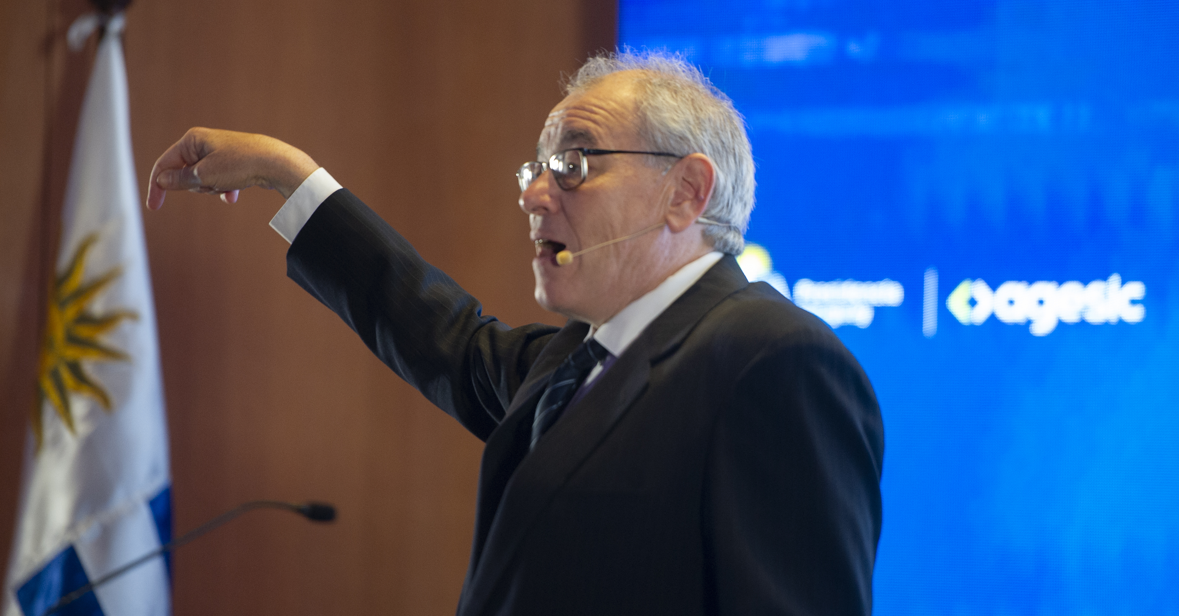 Director de Agesic de pie con micrófono de vincha gesticula mientras diserta en el escenario. Atrás se observa el pabellón nacional.