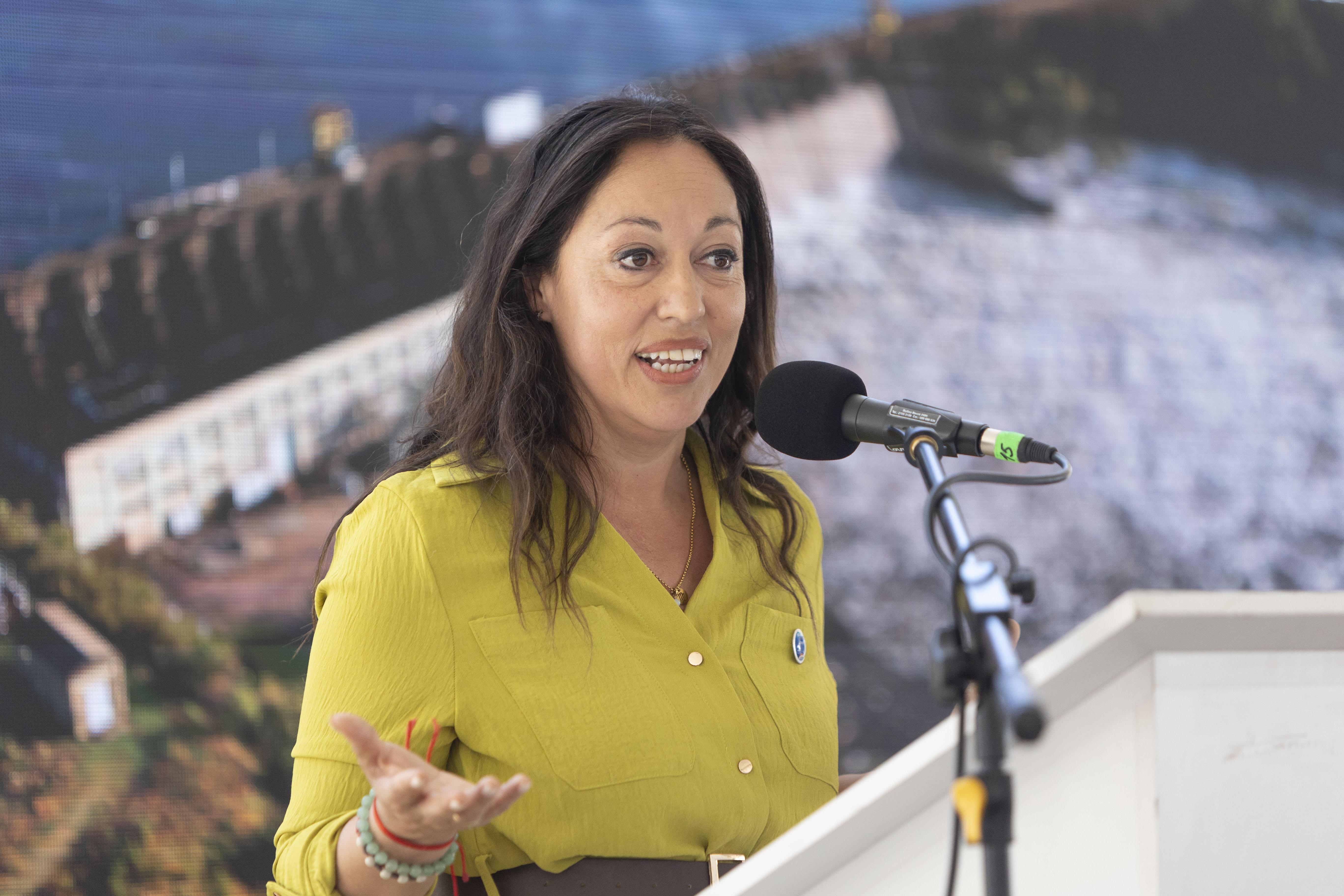 Ministra de Industria diserta de pie en atril con micrófono. Atrás se observa una gran imagen de la central hidroeléctrica.
