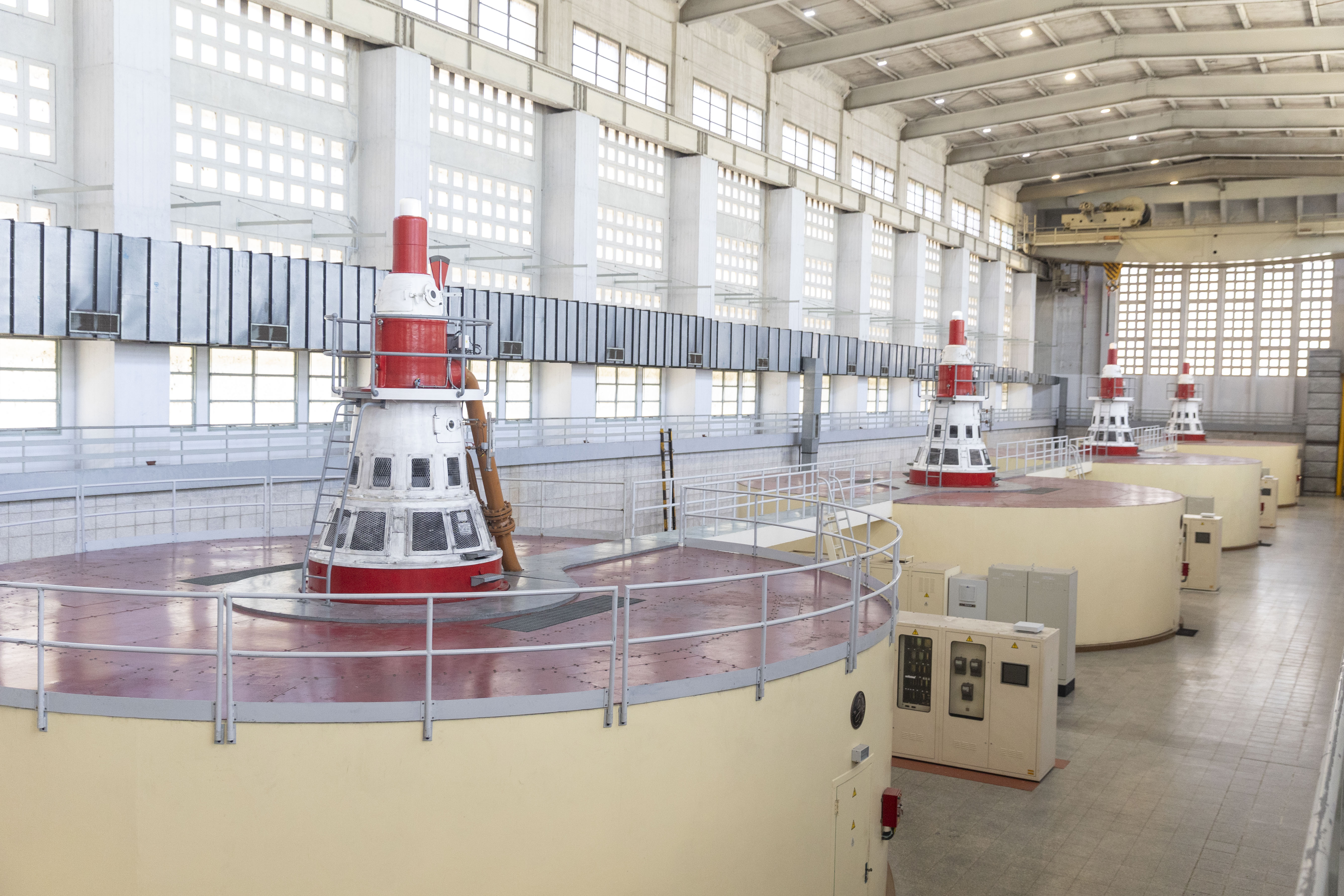 Interior de una central hidroeléctrica con varias unidades generadoras. Grandes ventanales en la estructura y techos altos.