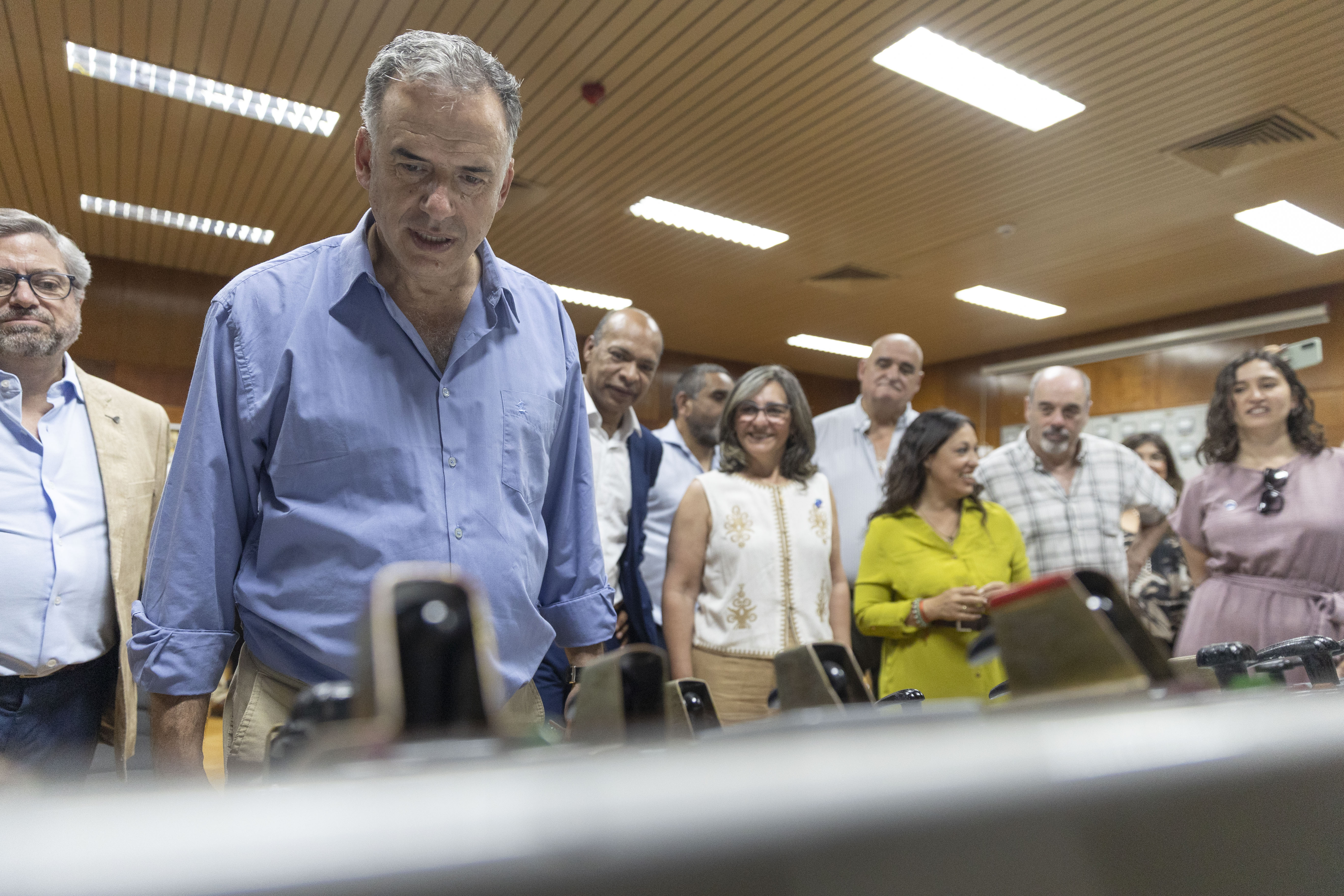 Orsi mira atentamente un panel de control en el interior de la central. Atrás varias personas lo observan.