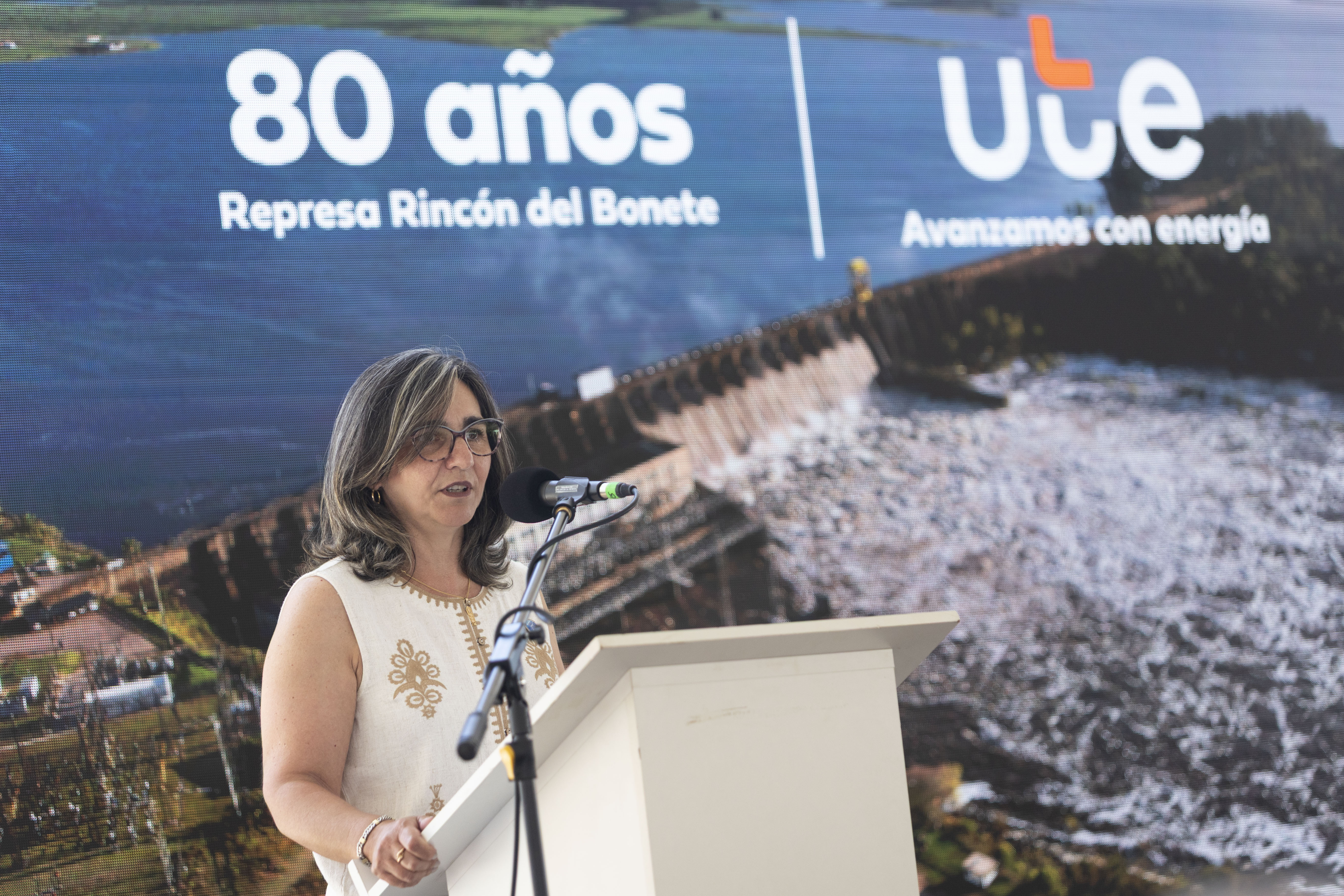 Presidenta de UTE diserta de pie en atril con micrófono. Atrás se observa gran cartel con foto de la represa y el logo de UTE.