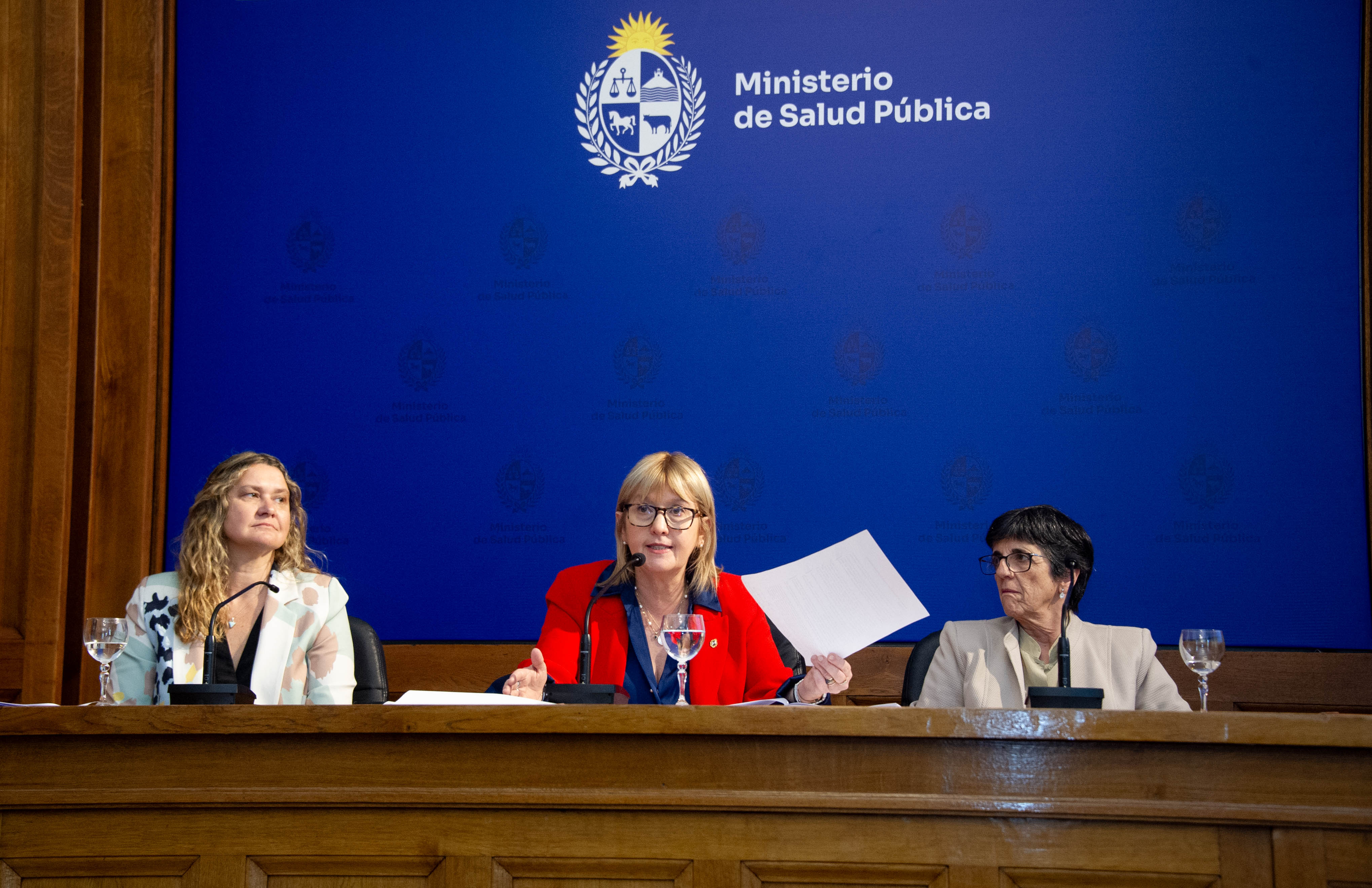 Ministra de Salud Pública diserta sentada en mesa con micrófono con un documento en una de sus manos. A sus costados están las otras autoridades.